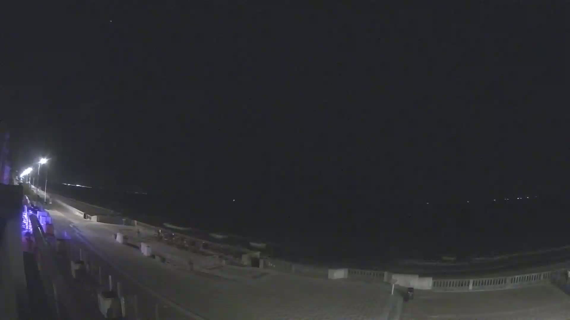 A nighttime view of a mostly empty beach boardwalk and ocean under a clear, dark sky.