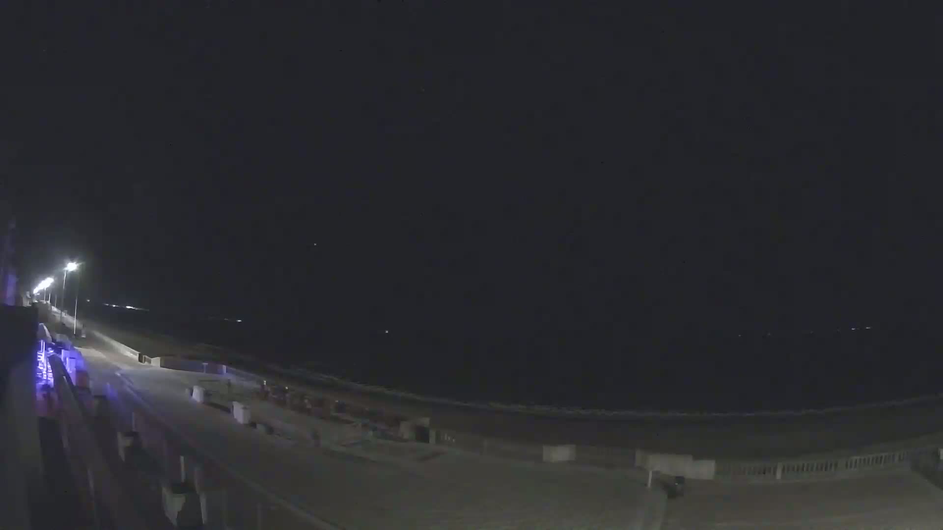 A nighttime view shows a dark beach with a boardwalk extending to the left, lit by streetlights, under a clear, dark sky.