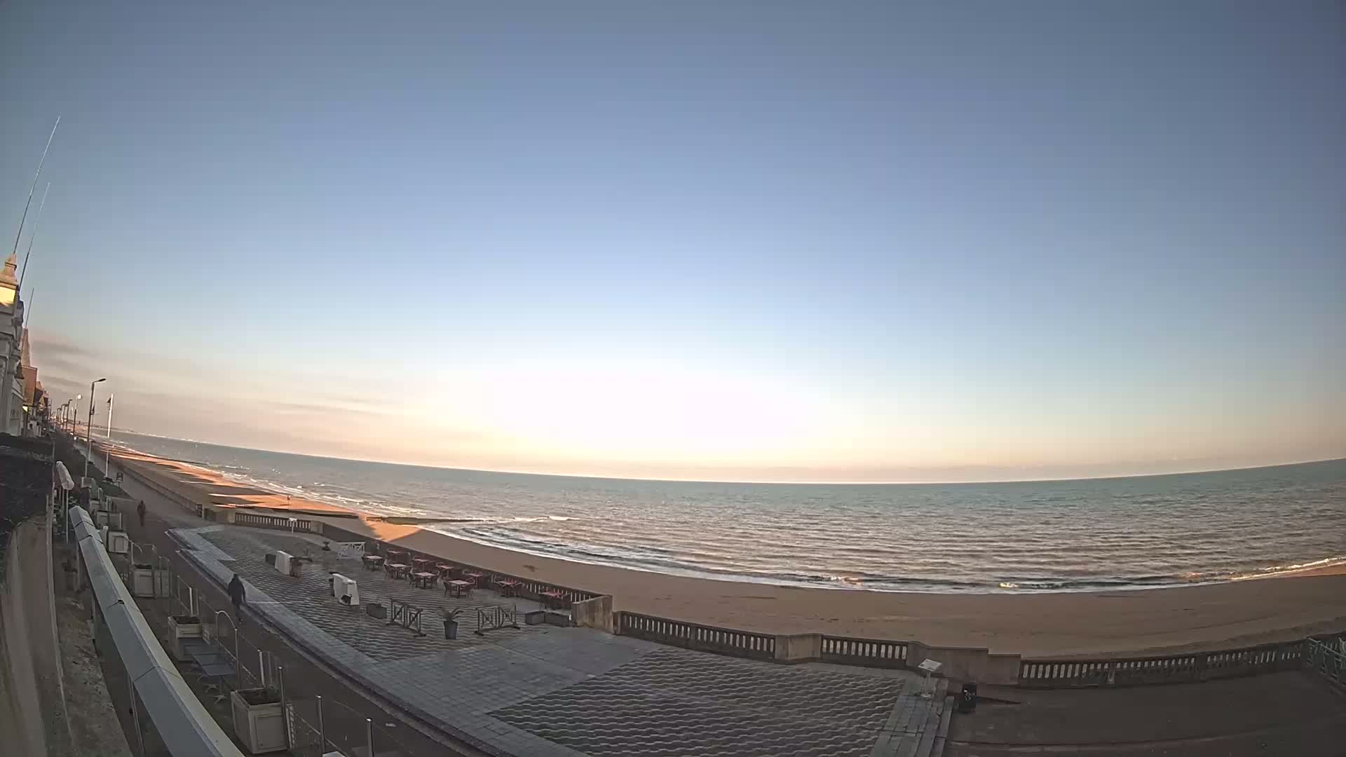 A calm ocean with a sandy beach is seen from a seaside promenade under a clear, bright sky.