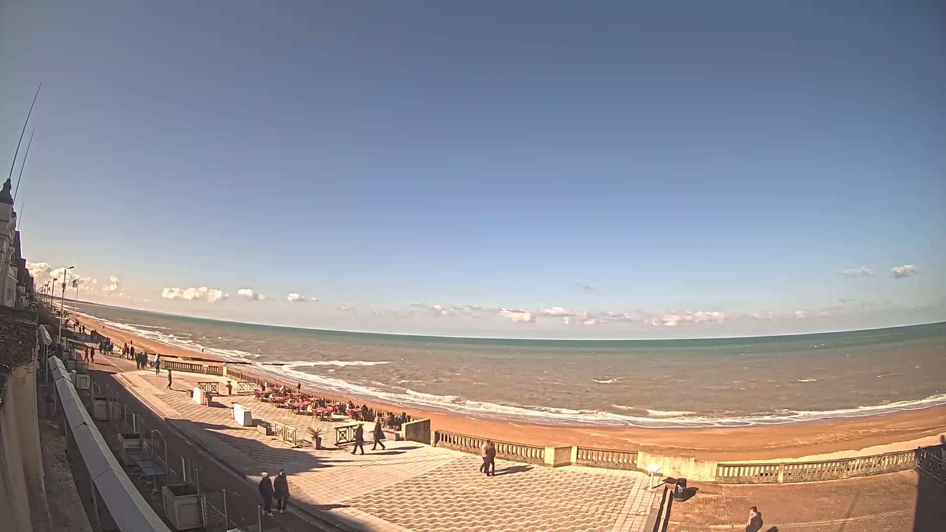 A mostly sunny day shows a wide expanse of ocean meeting a sandy beach, with a paved walkway and a few people visible along the shore.
