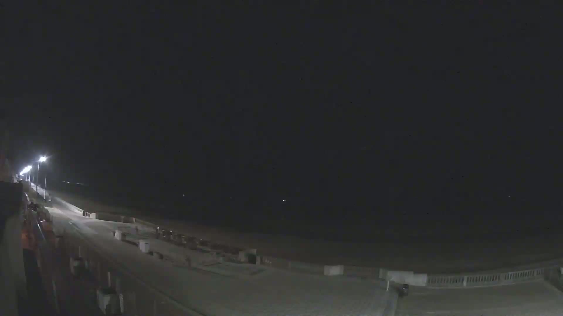 A nighttime view of a mostly empty beach boardwalk with a few lights illuminating the path and dark skies above.