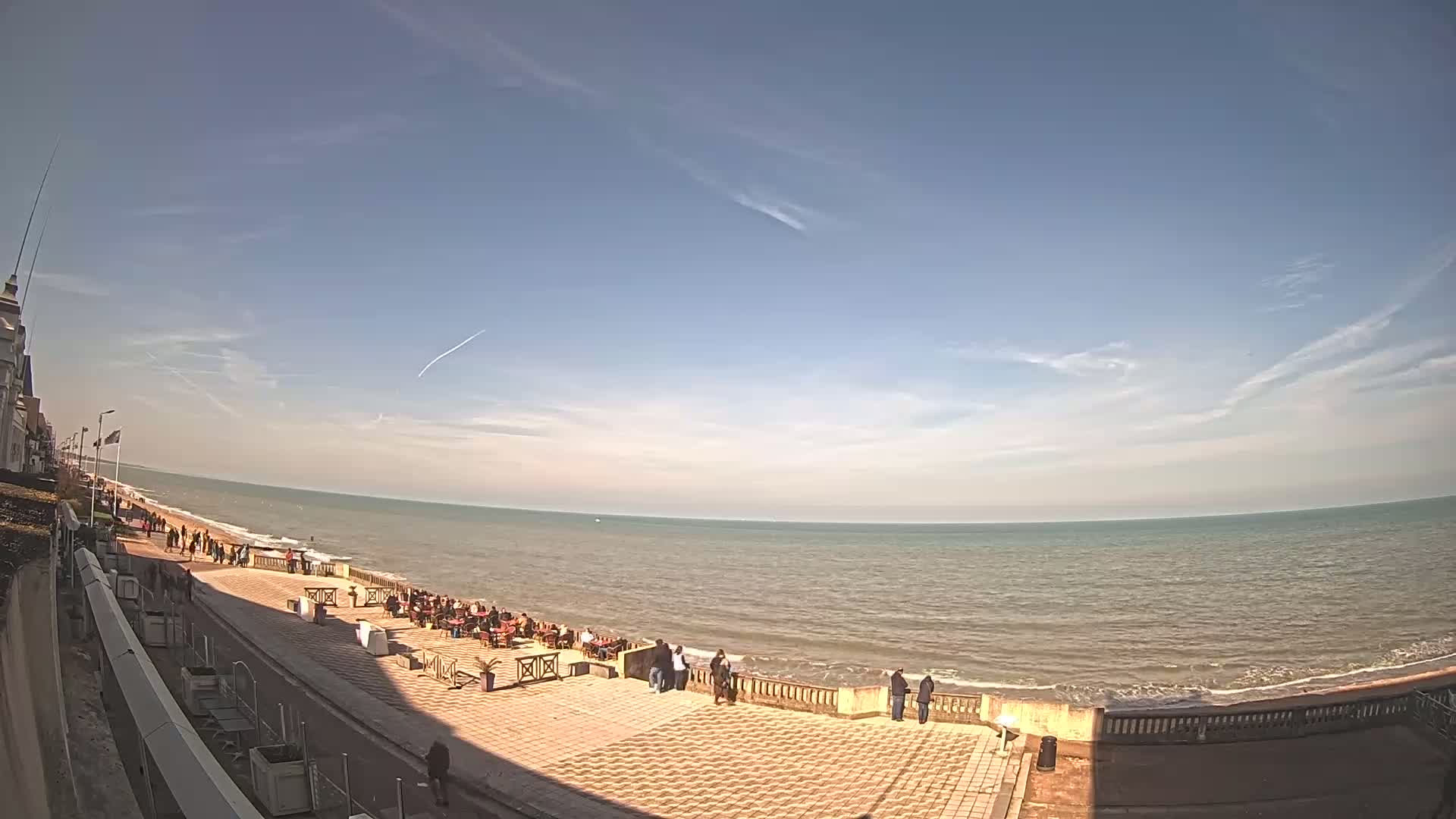 A sunny day shows a wide expanse of calm ocean meeting a paved beachfront promenade with people walking and sitting at outdoor cafes.