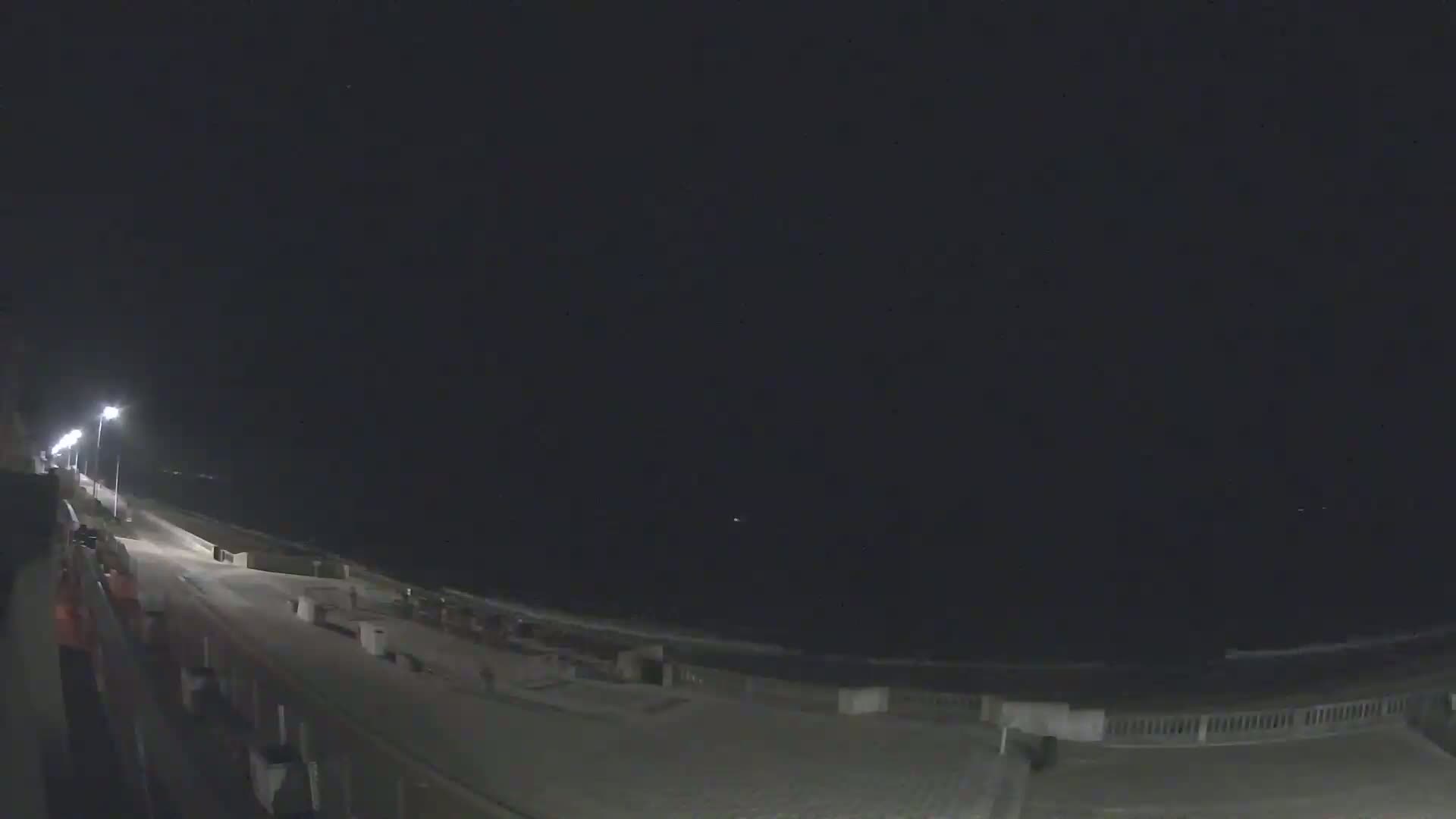 A nighttime view of a mostly empty beach boardwalk with streetlights illuminating the path and ocean waves visible in the distance under a dark sky.