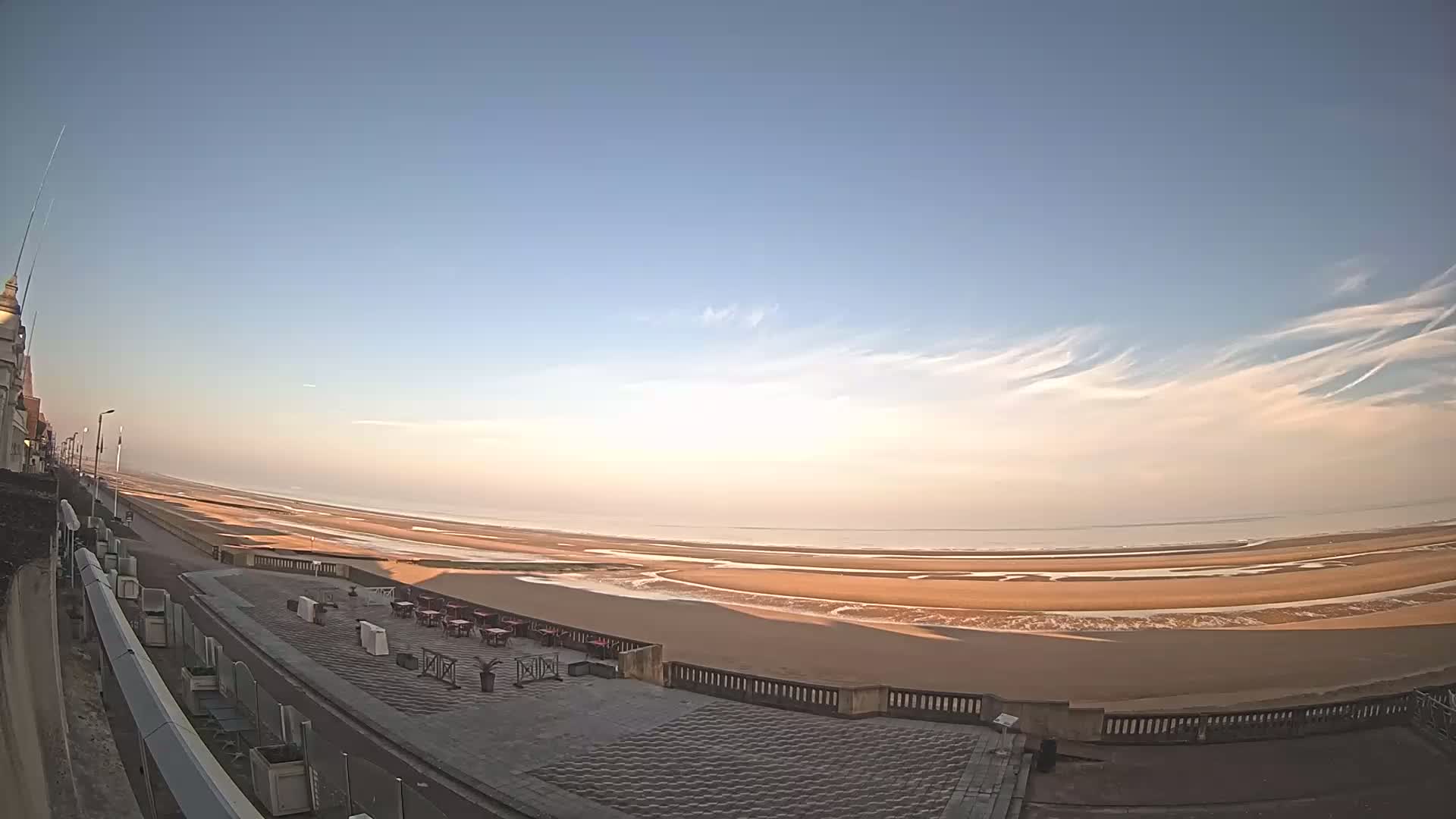 A wide, sandy beach stretches to a calm ocean under a clear, pale blue sky with thin clouds.