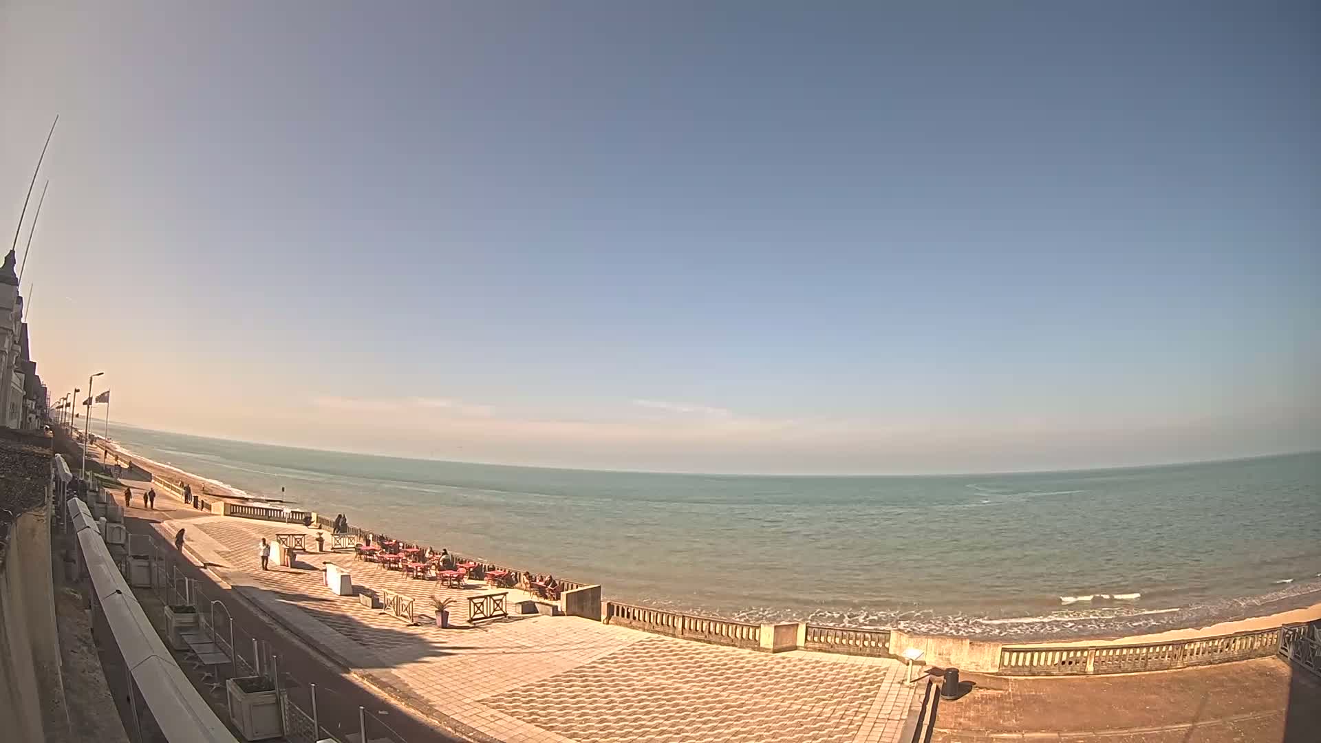 A paved beachfront area with a few people and outdoor seating overlooks a calm ocean under a clear blue sky.