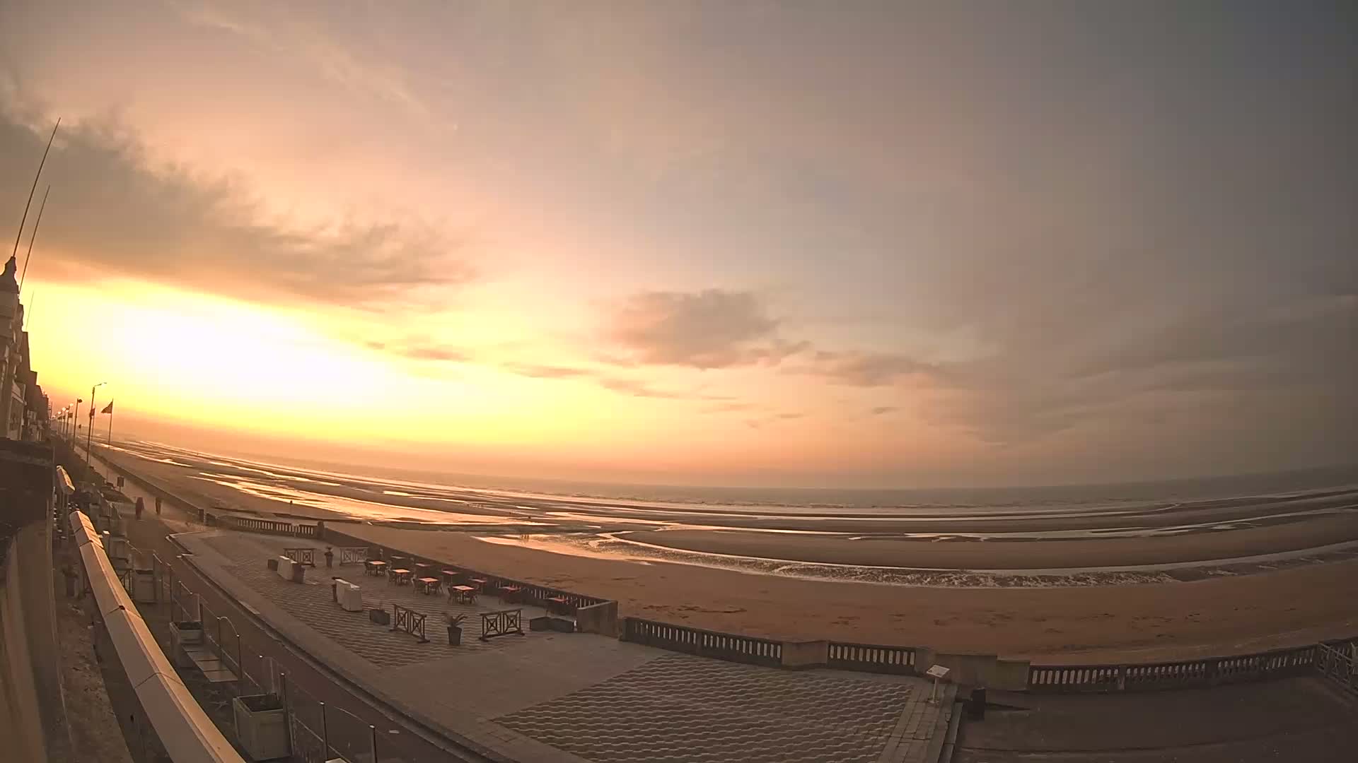 A wide shot shows a sandy beach at sunset under a partly cloudy sky, with a paved walkway and outdoor seating area visible near a building in the foreground.