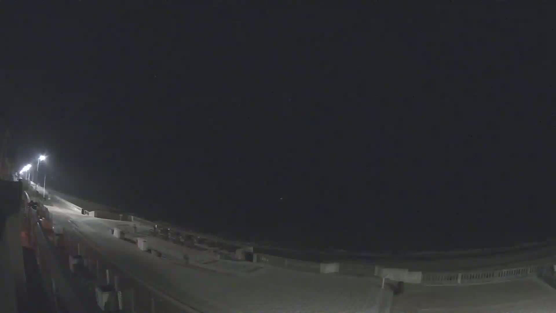 A nighttime, dimly lit beachfront scene shows a paved walkway leading to the ocean under a dark sky.