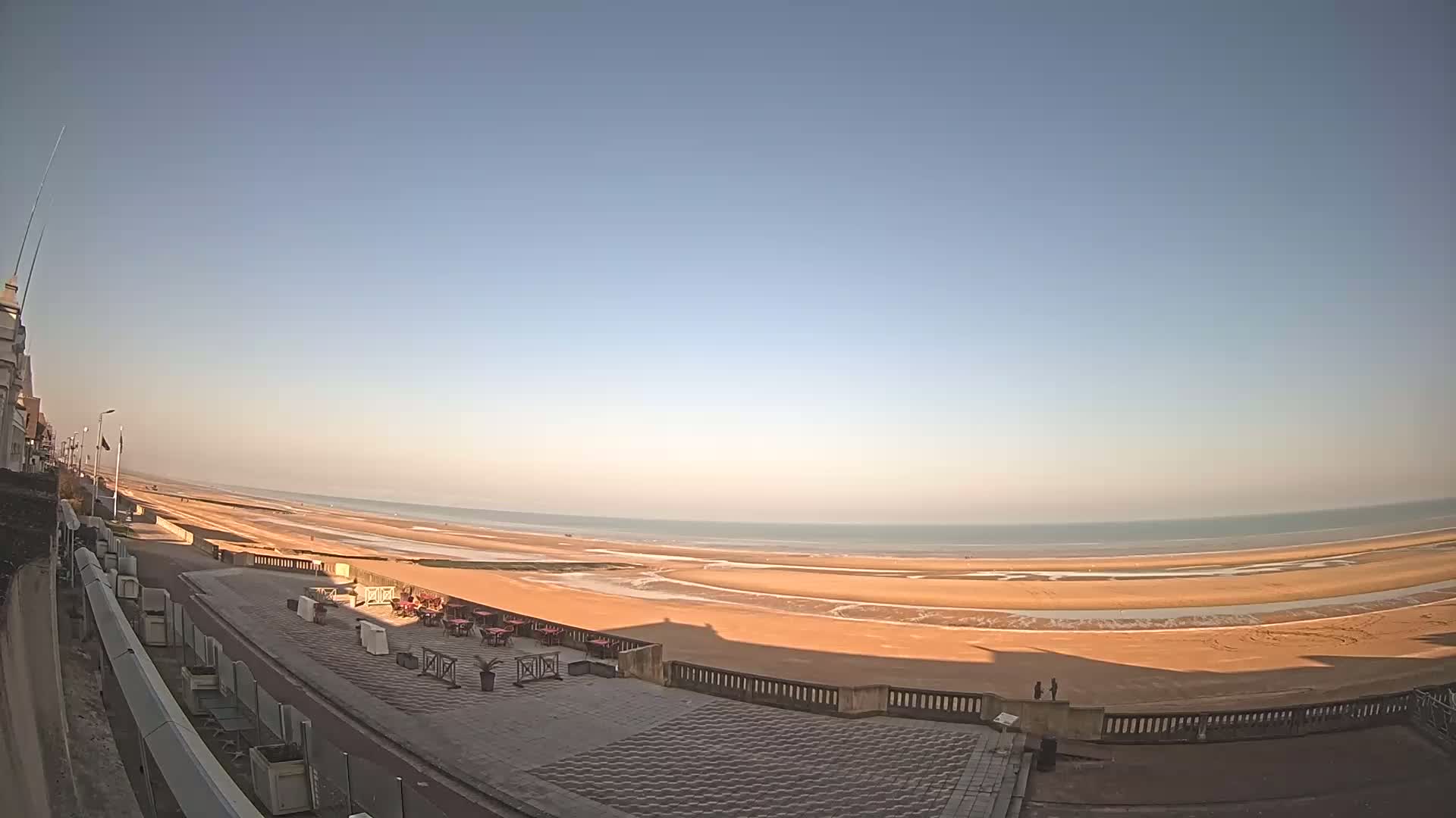 A wide, sandy beach stretches to a calm ocean under a clear blue sky, with a paved promenade and outdoor seating area in the foreground.