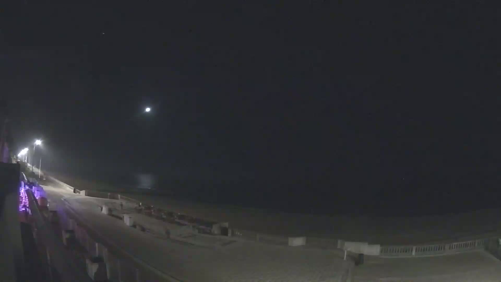 A nighttime view of a beach with a boardwalk, lit by streetlights under a dark, mostly clear sky.