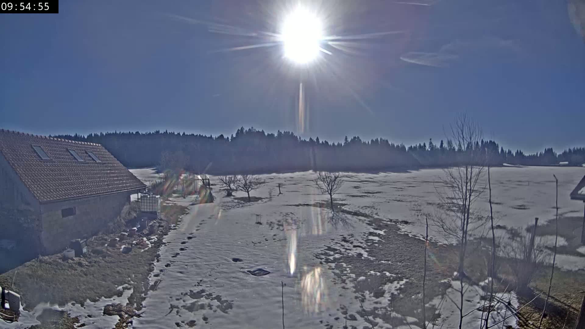 A snow-covered field stretches behind a stone building under a bright, sunny sky.