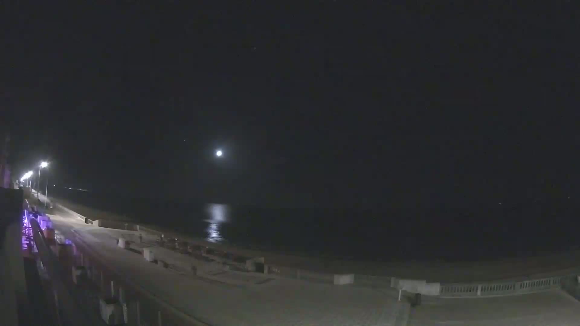 A moonlit nighttime scene shows a paved beachfront area with a calm ocean and streetlights lining a walkway.