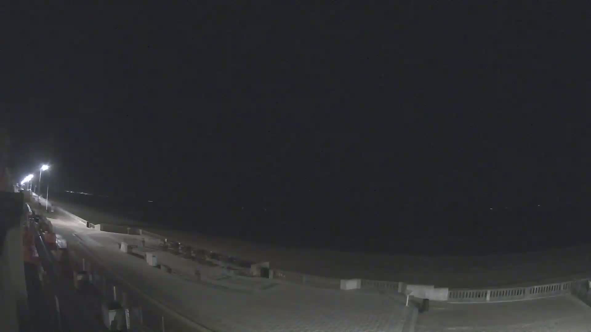 A nighttime view of a mostly empty beach boardwalk with lights illuminating the path and a dark ocean in the background under a clear sky.