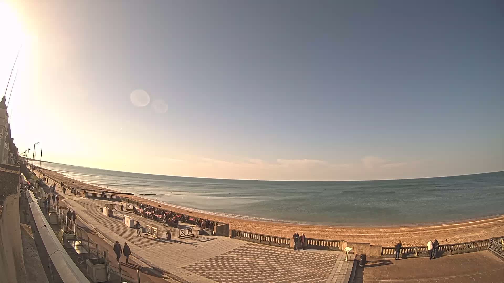 A sunny day at a beach shows a paved promenade with people walking and sitting at outdoor cafes, overlooking a calm ocean with a sandy shore.