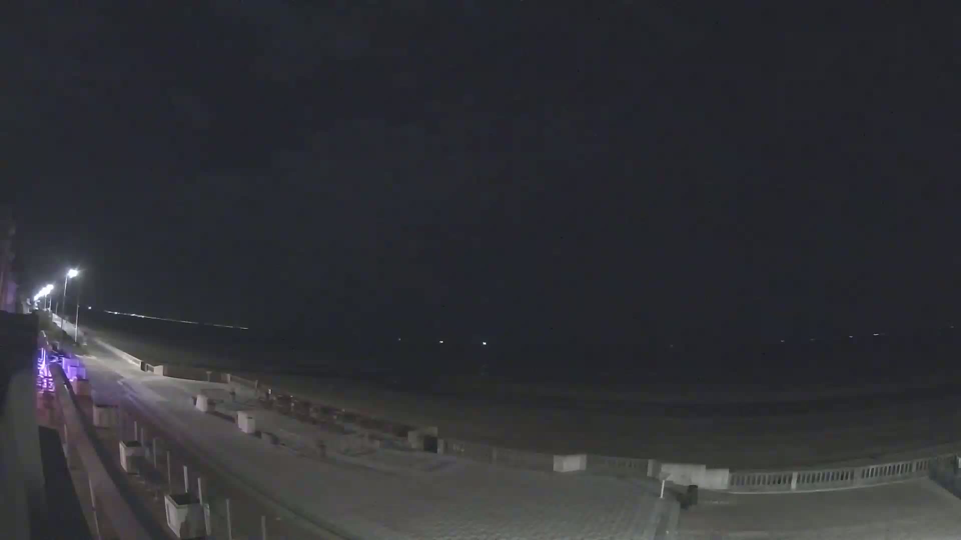 A nighttime view shows a dark beach with a paved walkway and low lighting, under a clear, dark sky.