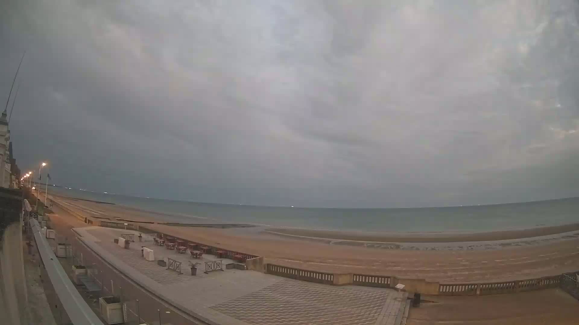 An overcast sky hangs over a calm sea and sandy beach, with an empty promenade and outdoor seating area visible in the foreground.