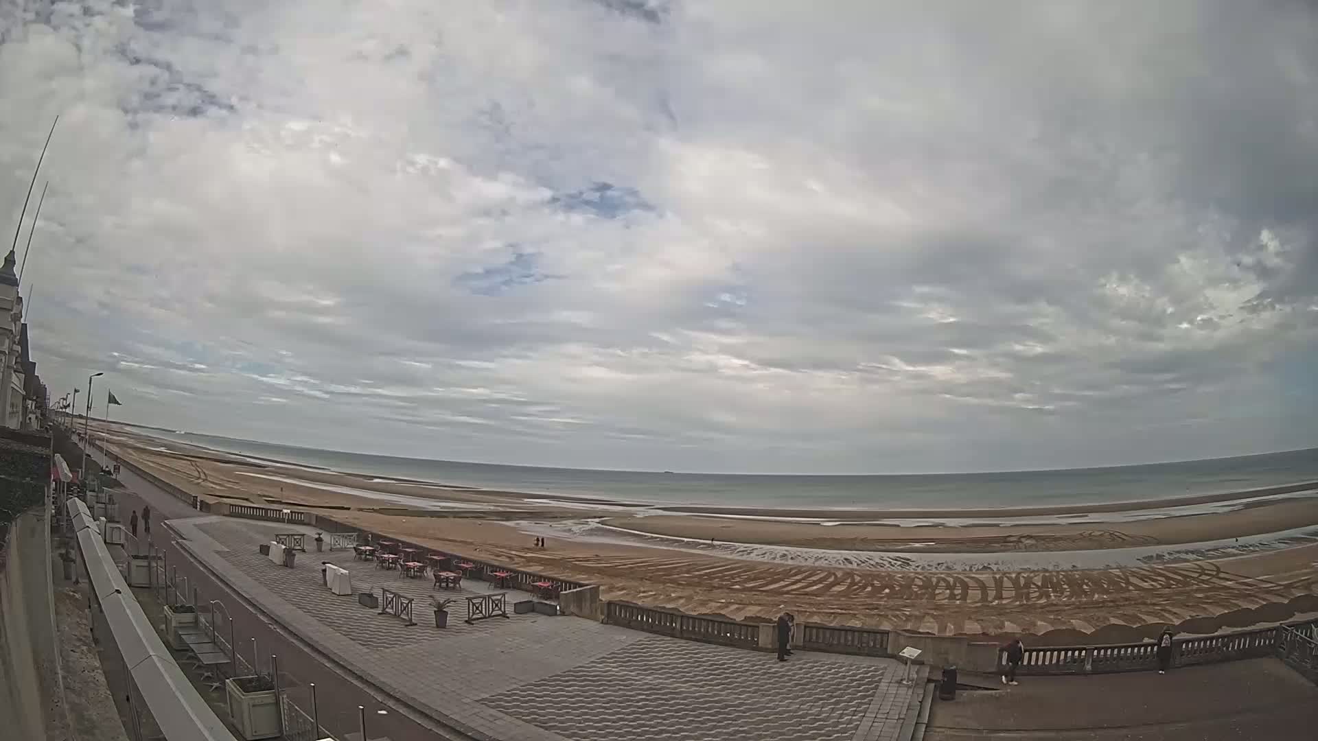 A wide, sandy beach stretches along a calm ocean under a mostly cloudy sky, with a paved promenade and outdoor seating area visible in the foreground.