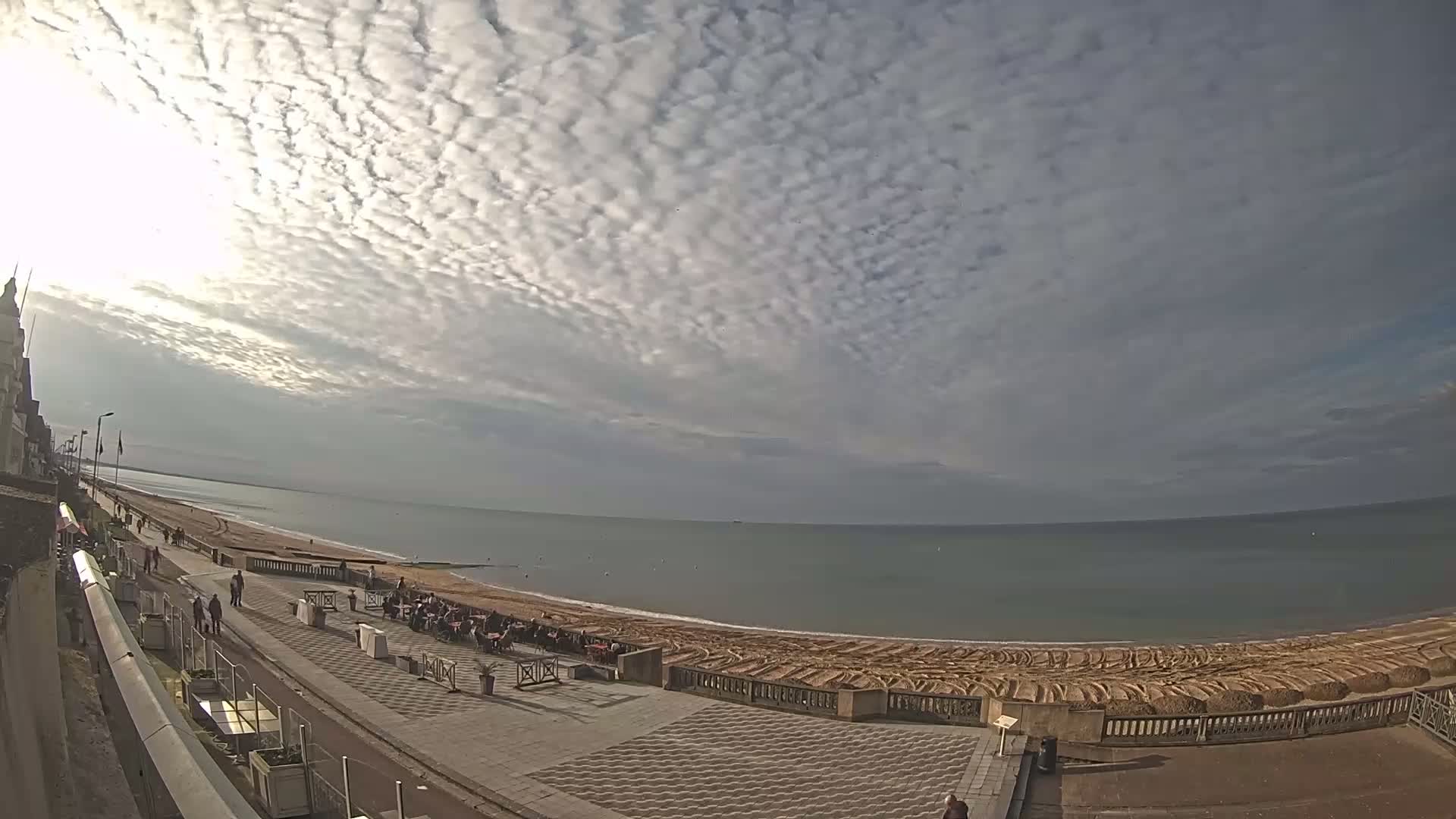 A mostly sunny day with a sky full of scattered, puffy clouds overlooks a beach with a paved walkway and people strolling along the shore.