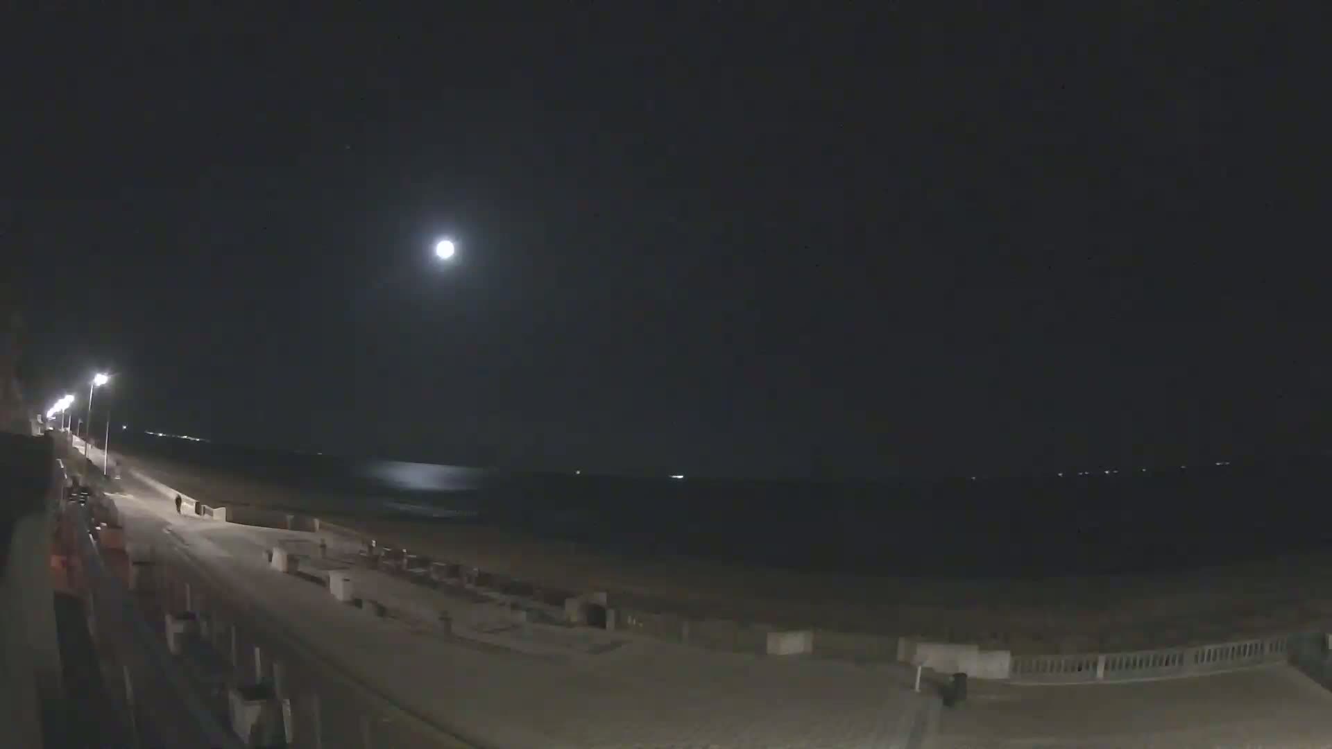 A brightly lit boardwalk overlooks a dark beach and ocean under a nighttime sky illuminated by a bright moon.