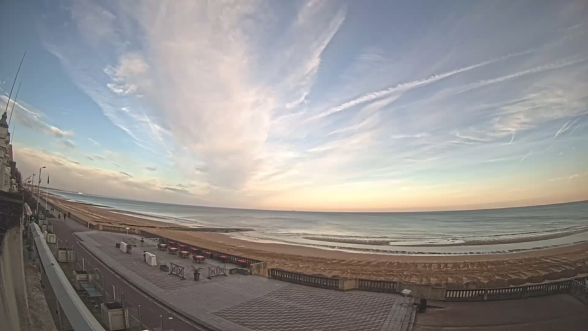 A wide shot of a beach with a paved walkway and outdoor seating area under a partly cloudy sky at sunset.