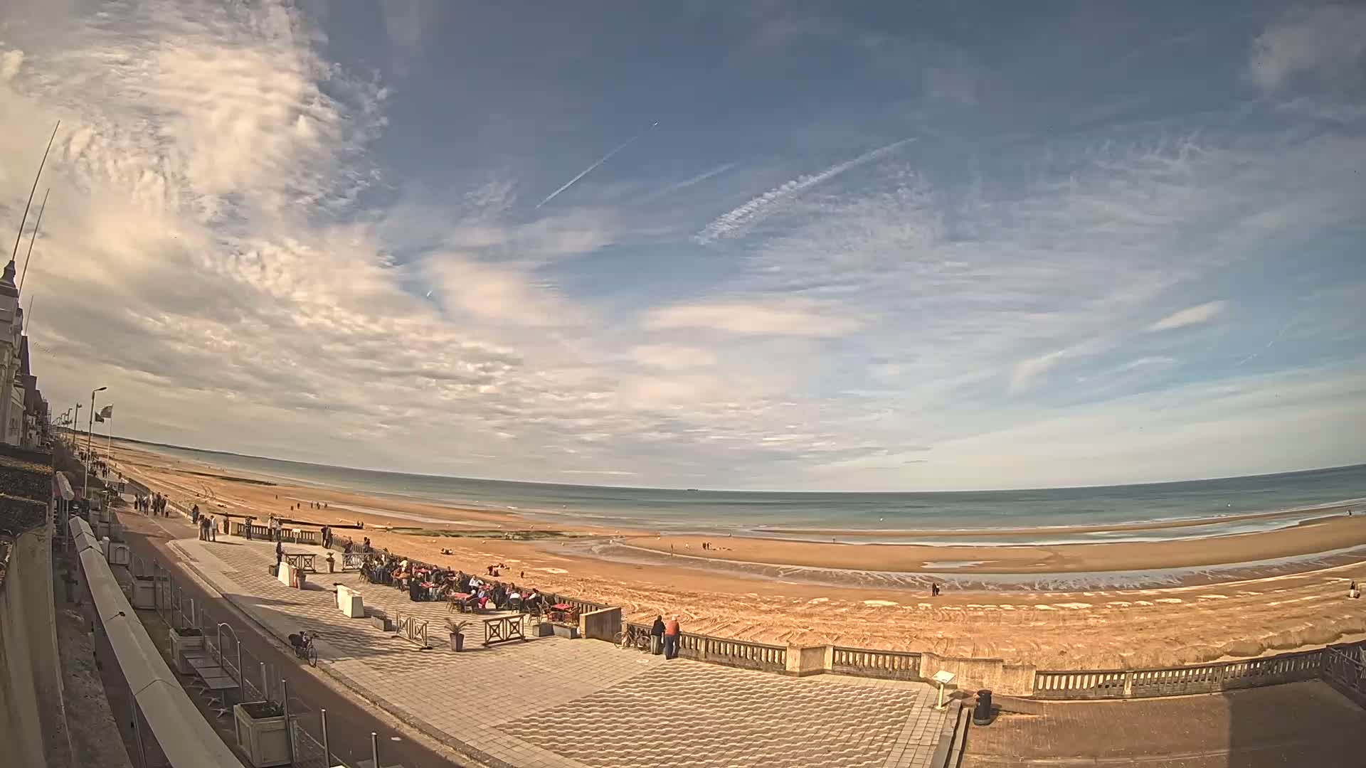 A wide-angle view shows a sandy beach and calm ocean under a mostly sunny sky with scattered clouds, overlooking a paved promenade with people and outdoor seating.