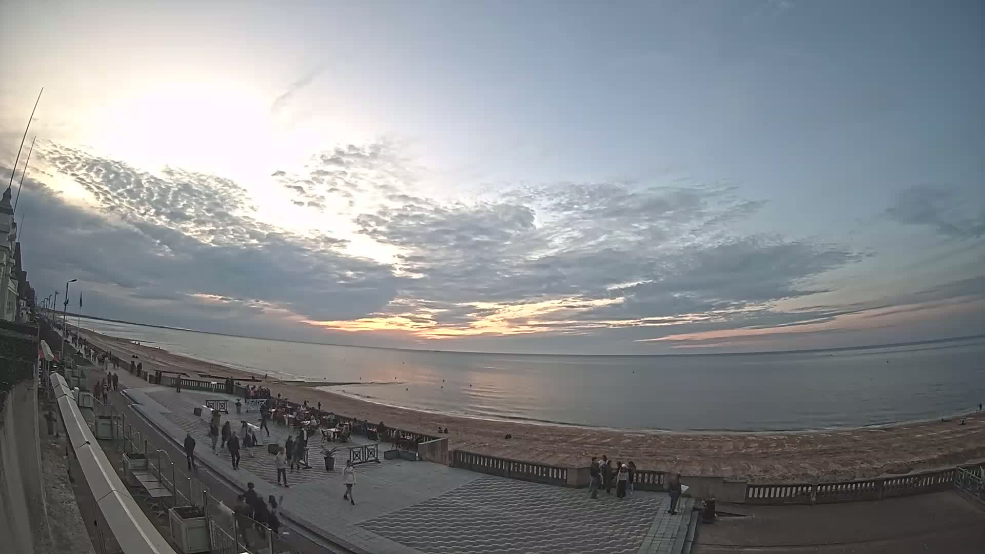 A wide shot shows a beach scene at sunset under a partly cloudy sky, with people walking along a promenade near the ocean.