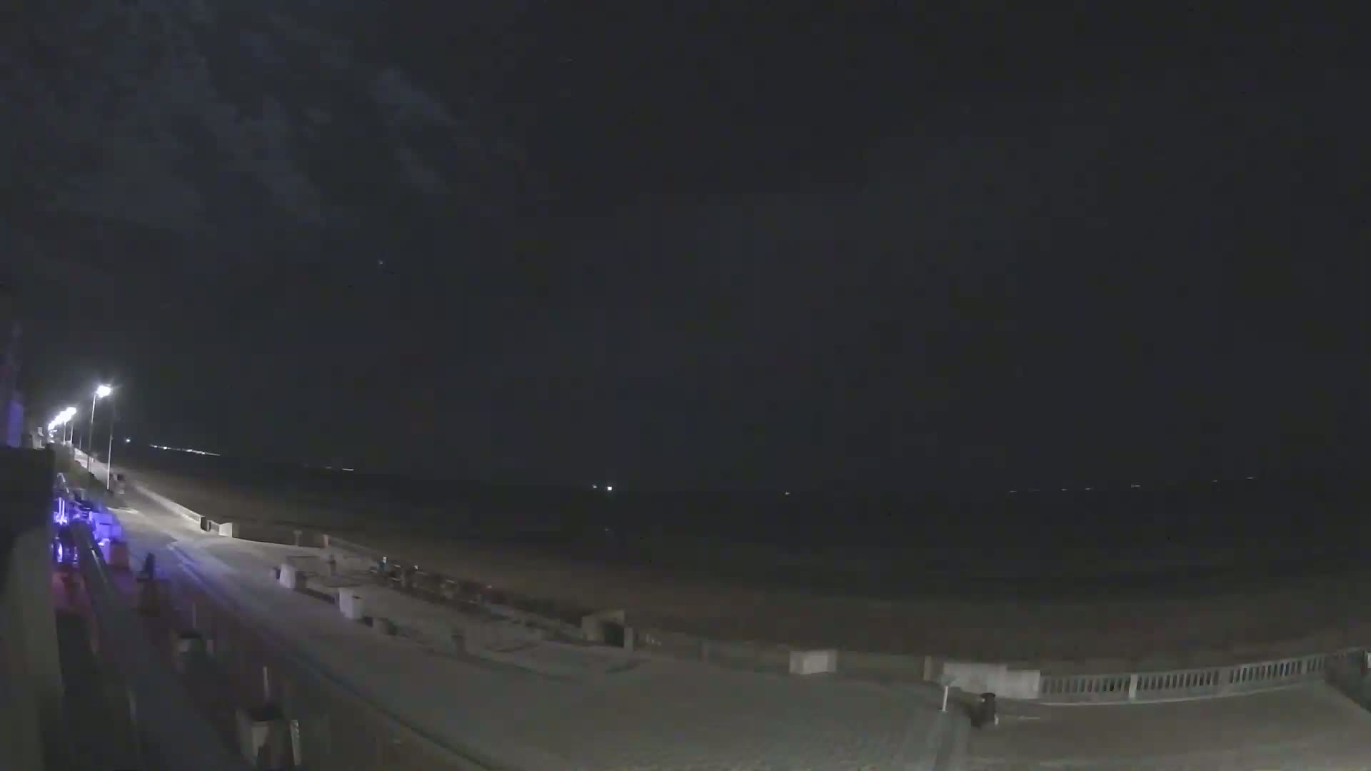 A nighttime view of a mostly empty beach boardwalk with a dark, cloudy sky and faintly visible lights across the water.