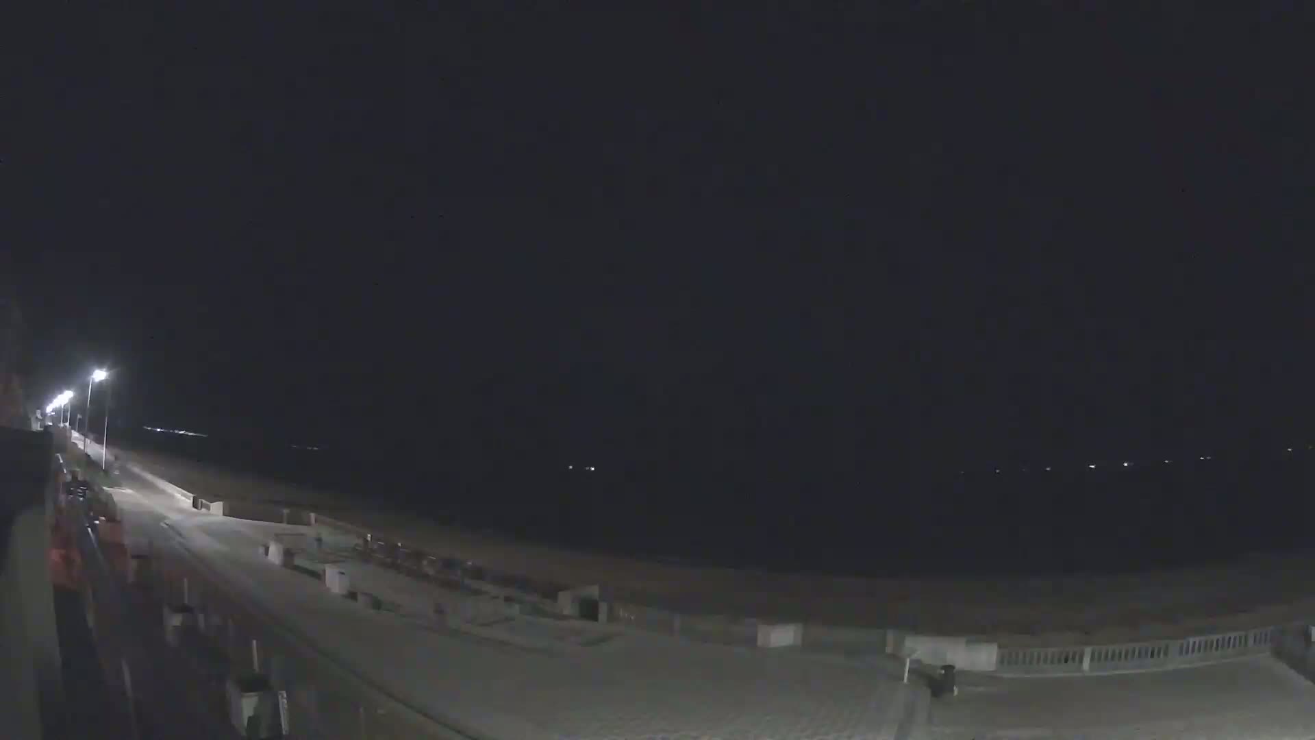 A nighttime view of a mostly empty beach boardwalk with the ocean visible in the distance under a clear, dark sky.