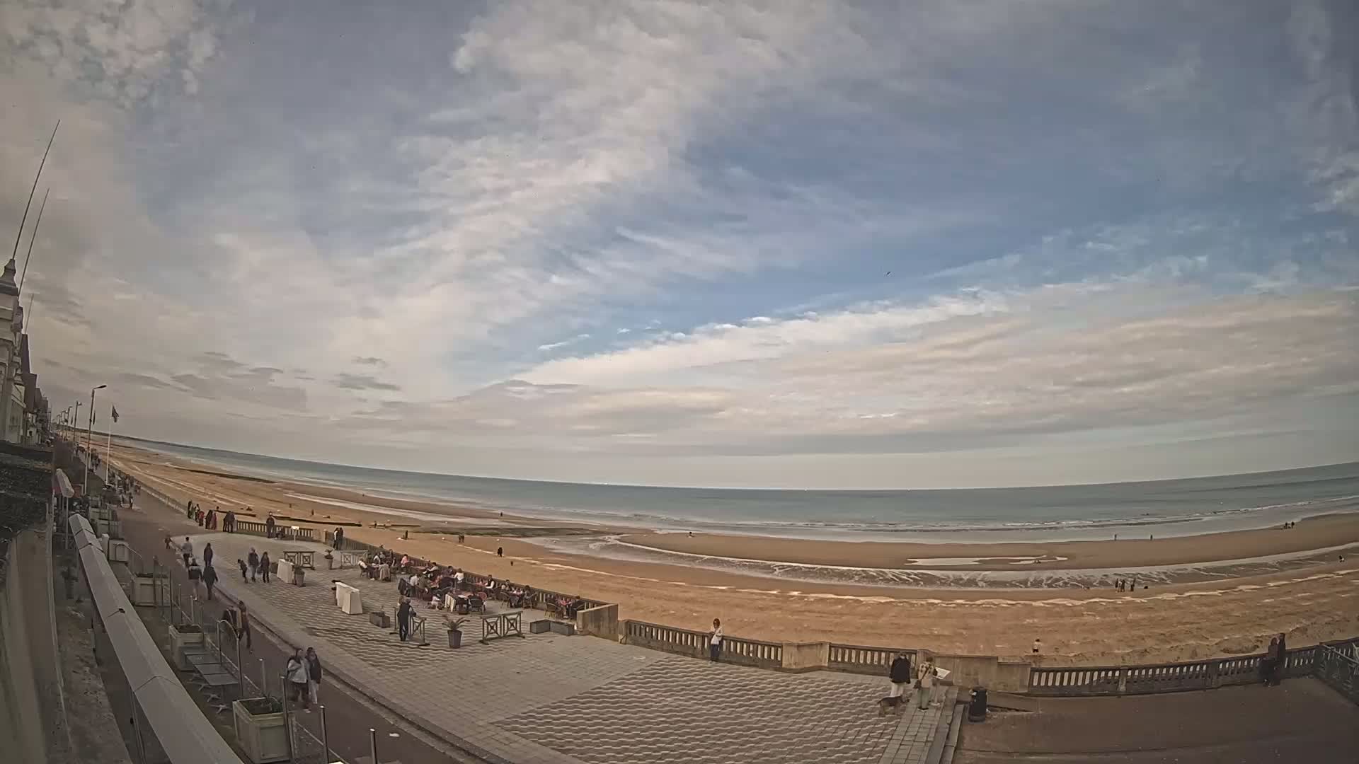 A wide shot shows a sandy beach with people walking along the shore and sitting at outdoor cafes under a partly cloudy sky.