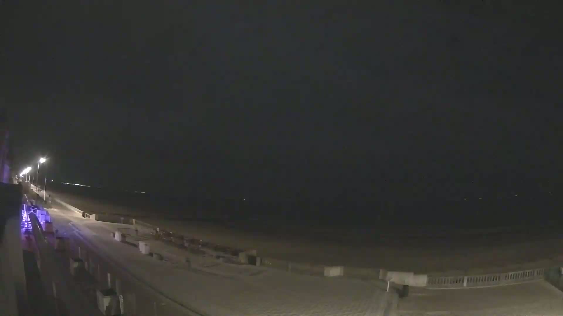 A dark, nighttime scene shows a snow-covered beach with a paved walkway and some lights along the edge.