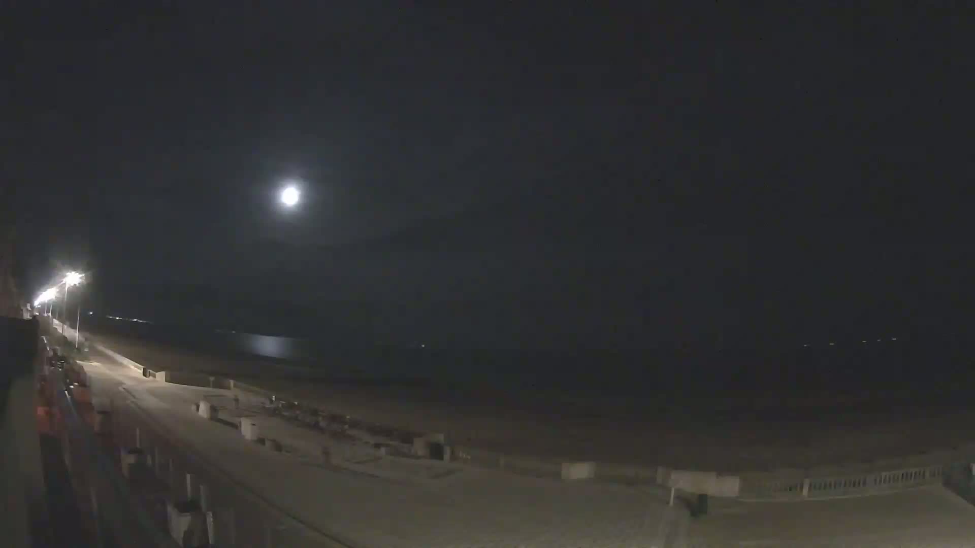A moonlit beach at night is seen from a high vantage point, showing a boardwalk and calm water.