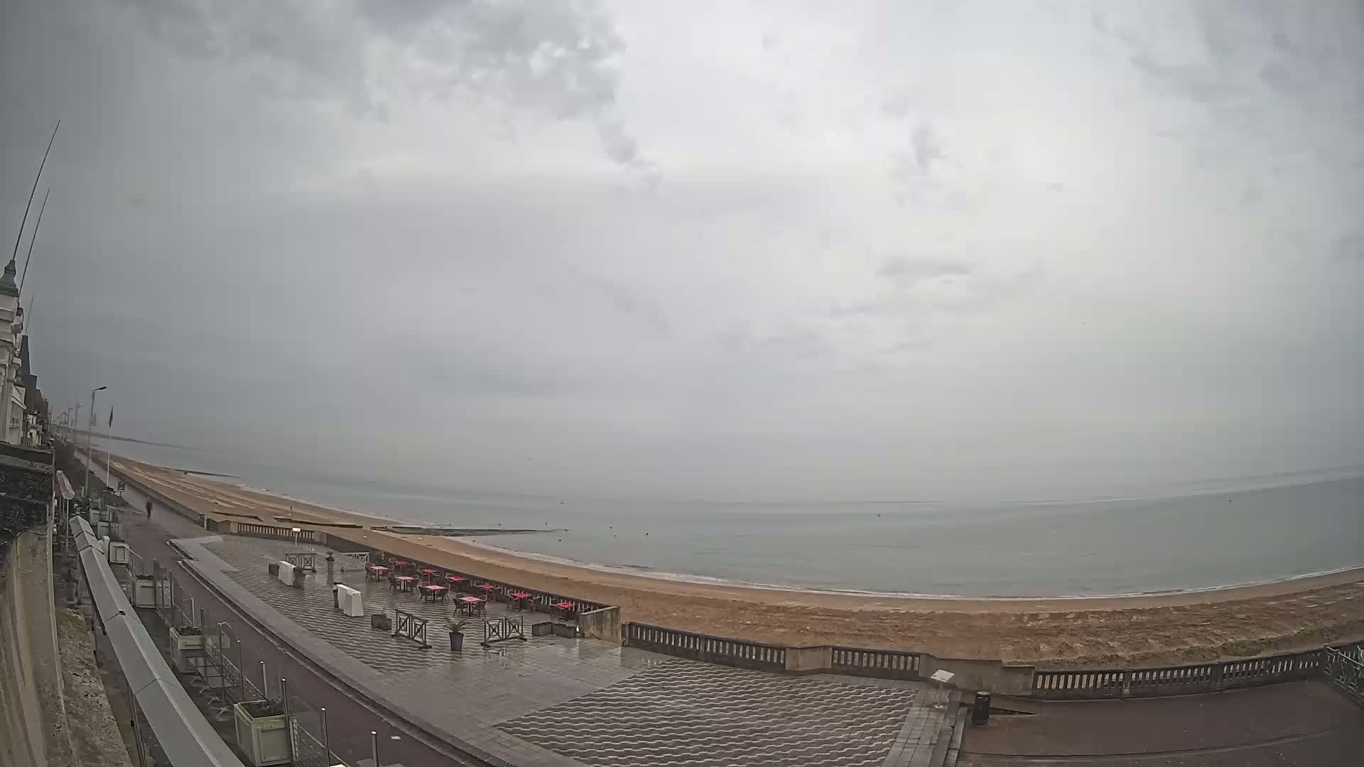 An overcast sky hangs over a mostly empty beachfront promenade with red tables and chairs set up near the sand.