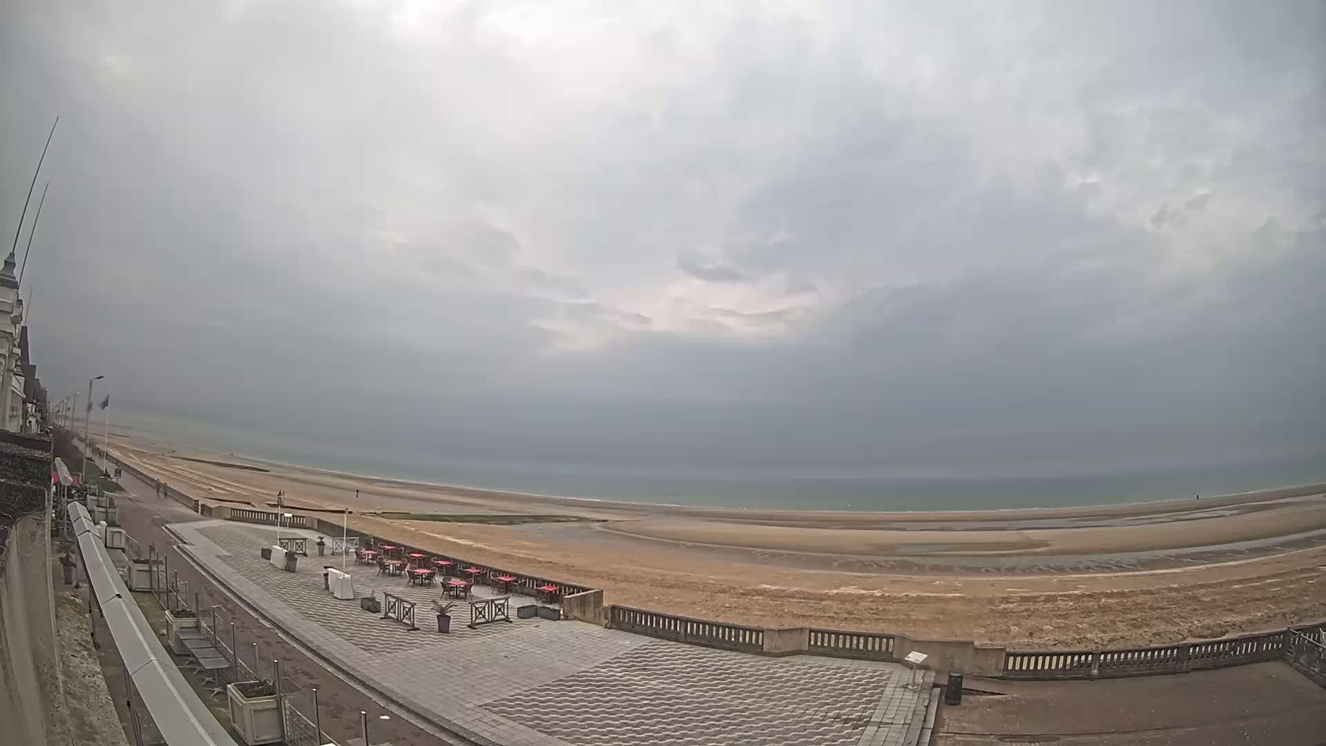 An overcast sky hangs over a wide, sandy beach and calm ocean, with an outdoor cafe area and a promenade visible in the foreground.