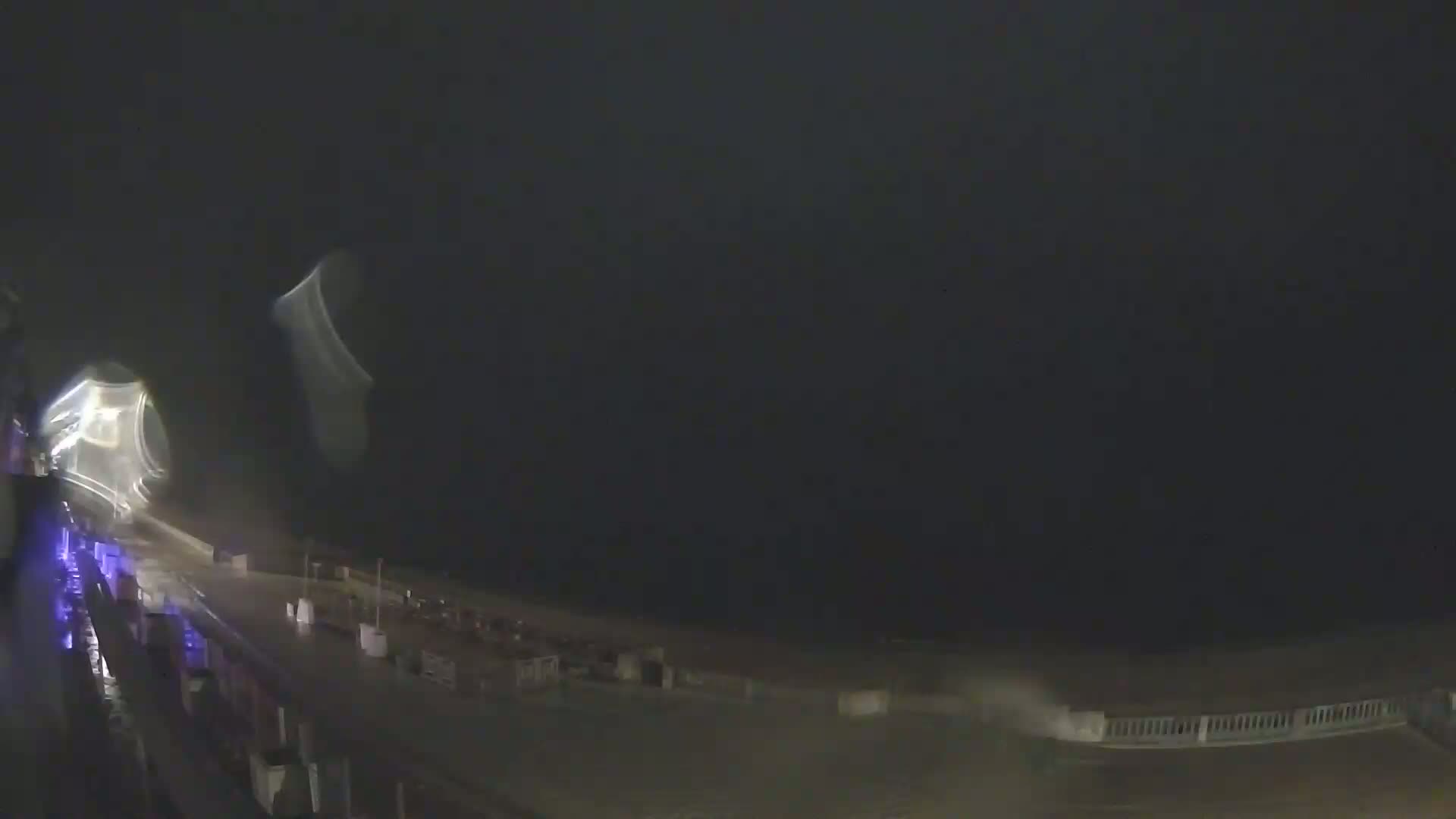 A nighttime, rain-swept view of a boardwalk overlooking a dark ocean under a stormy sky.