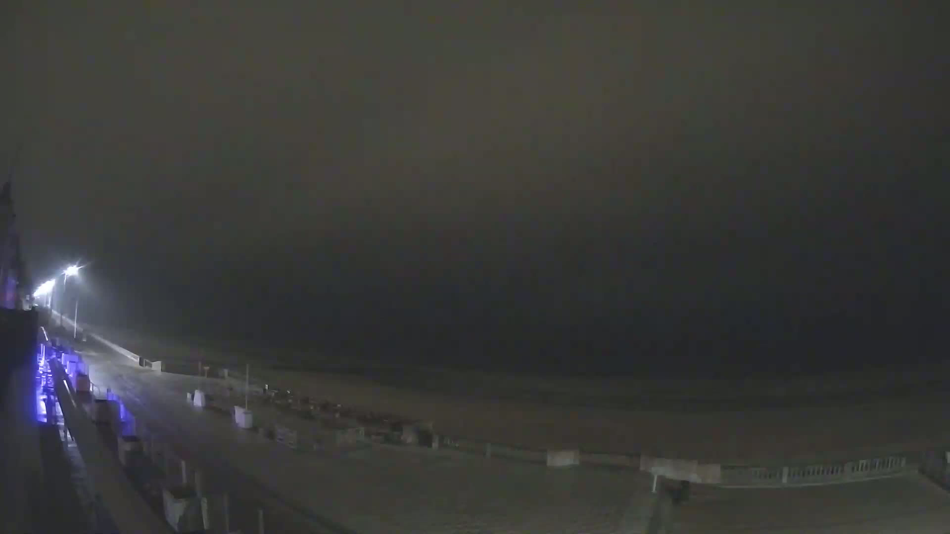 A nighttime, foggy view of a wet, empty beach with streetlights illuminating a roadway leading to it.
