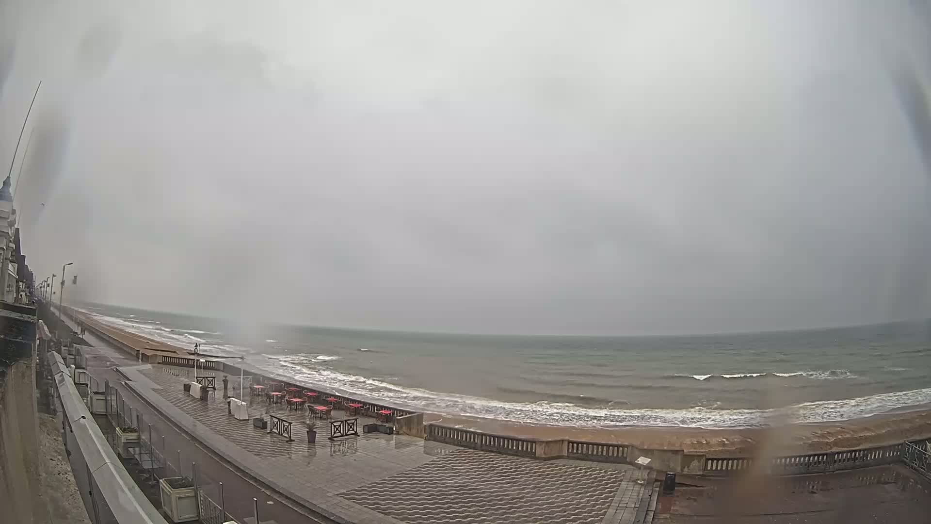 A rain-swept seaside promenade with an empty outdoor cafe overlooks a choppy sea under an overcast sky.
