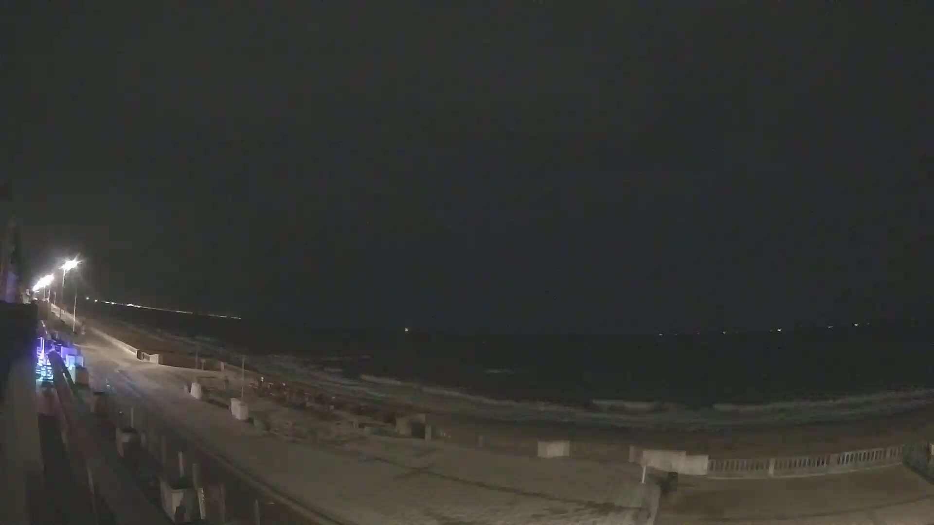 A nighttime view of a beach with gentle waves under a dark sky, lit by streetlights along a boardwalk.