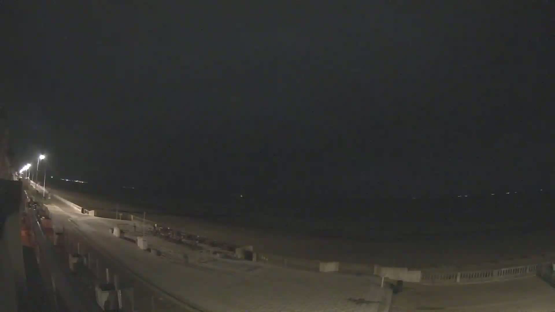 A nighttime view of a dimly lit beach boardwalk and shoreline under a dark, overcast sky.