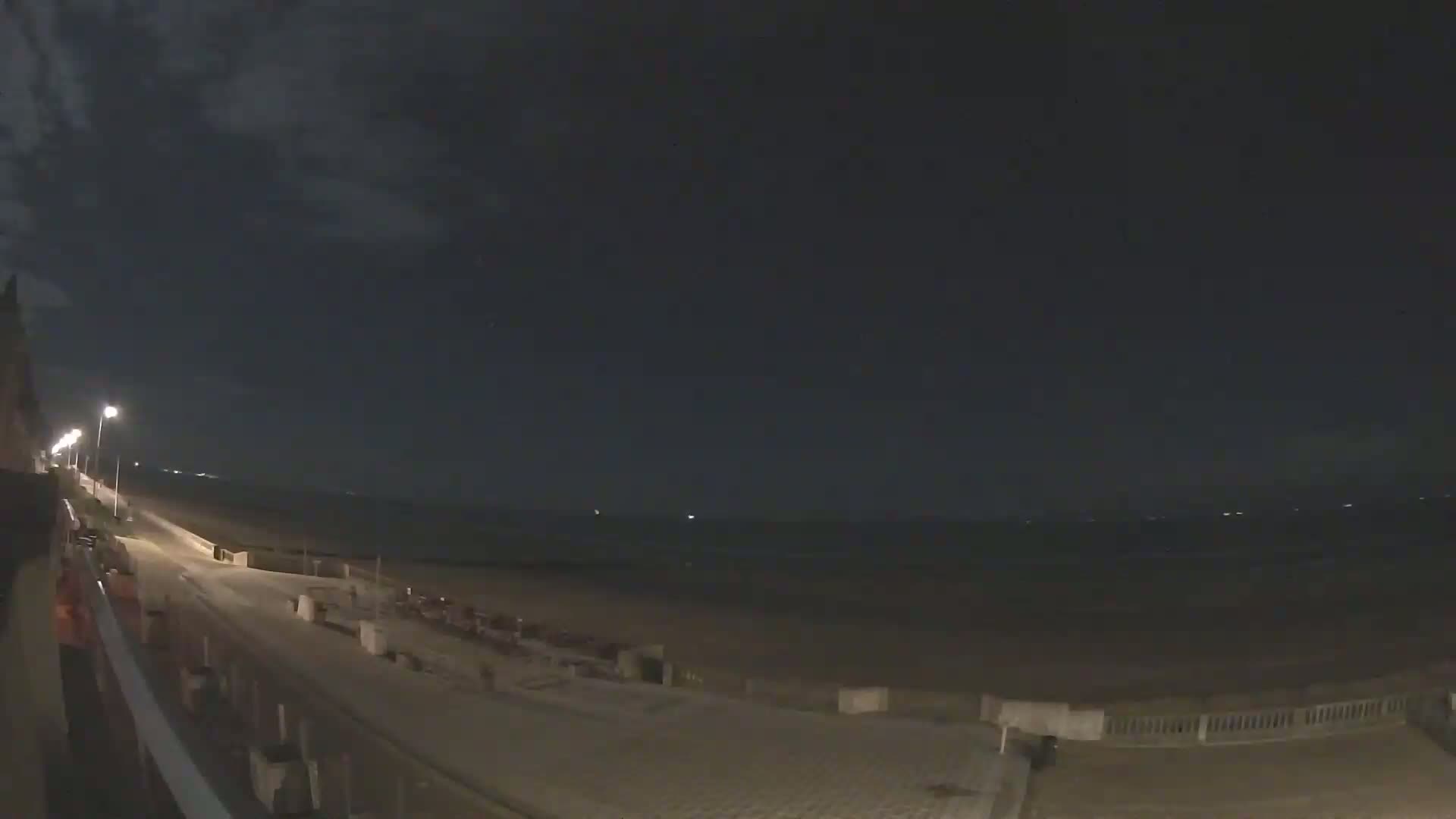 A nighttime view of a mostly empty beach boardwalk and sandy shore under a dark, cloudy sky.
