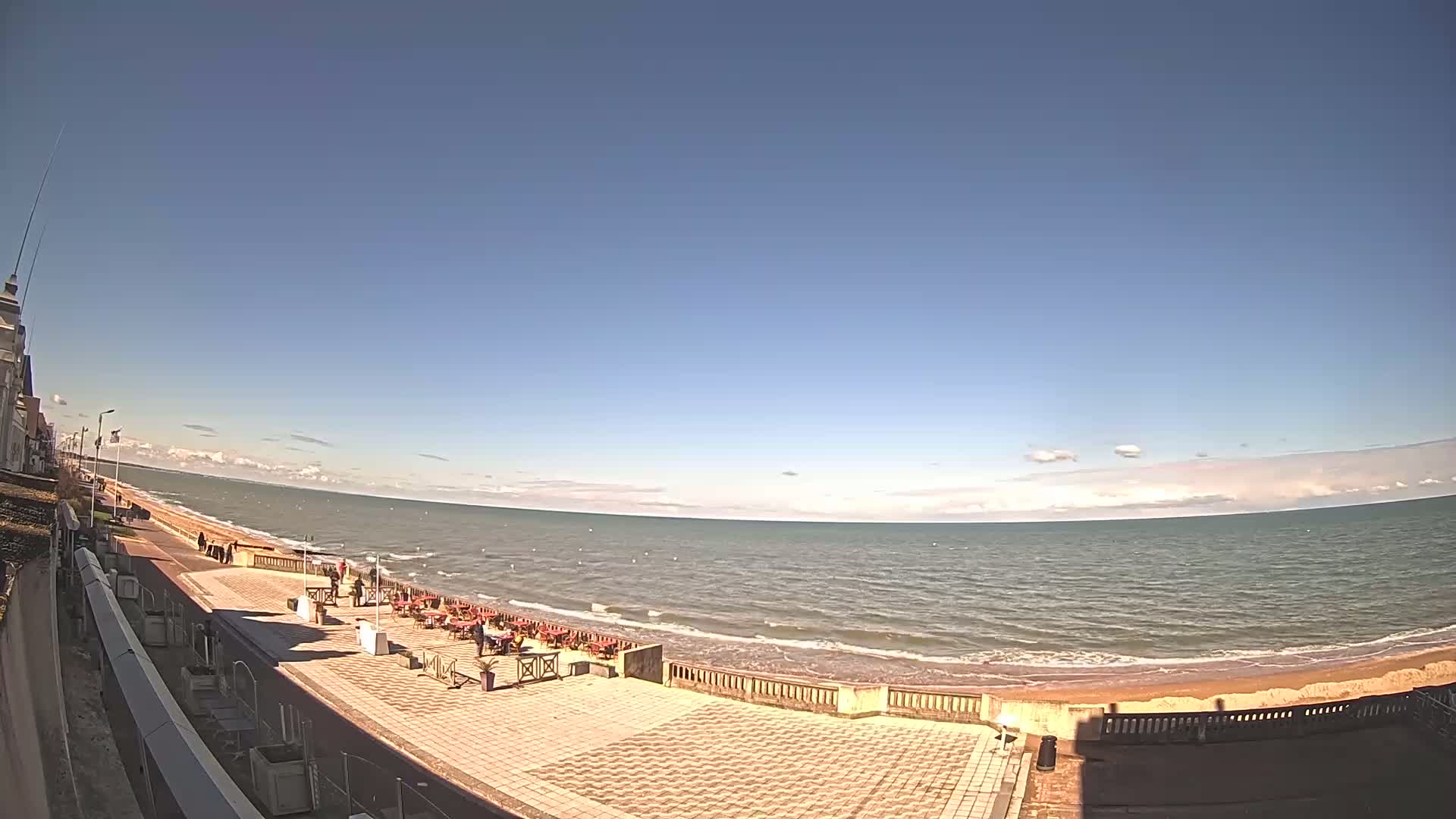 A paved beachfront promenade with a few people and outdoor seating overlooks a calm, light-blue ocean under a mostly clear, sunny sky.