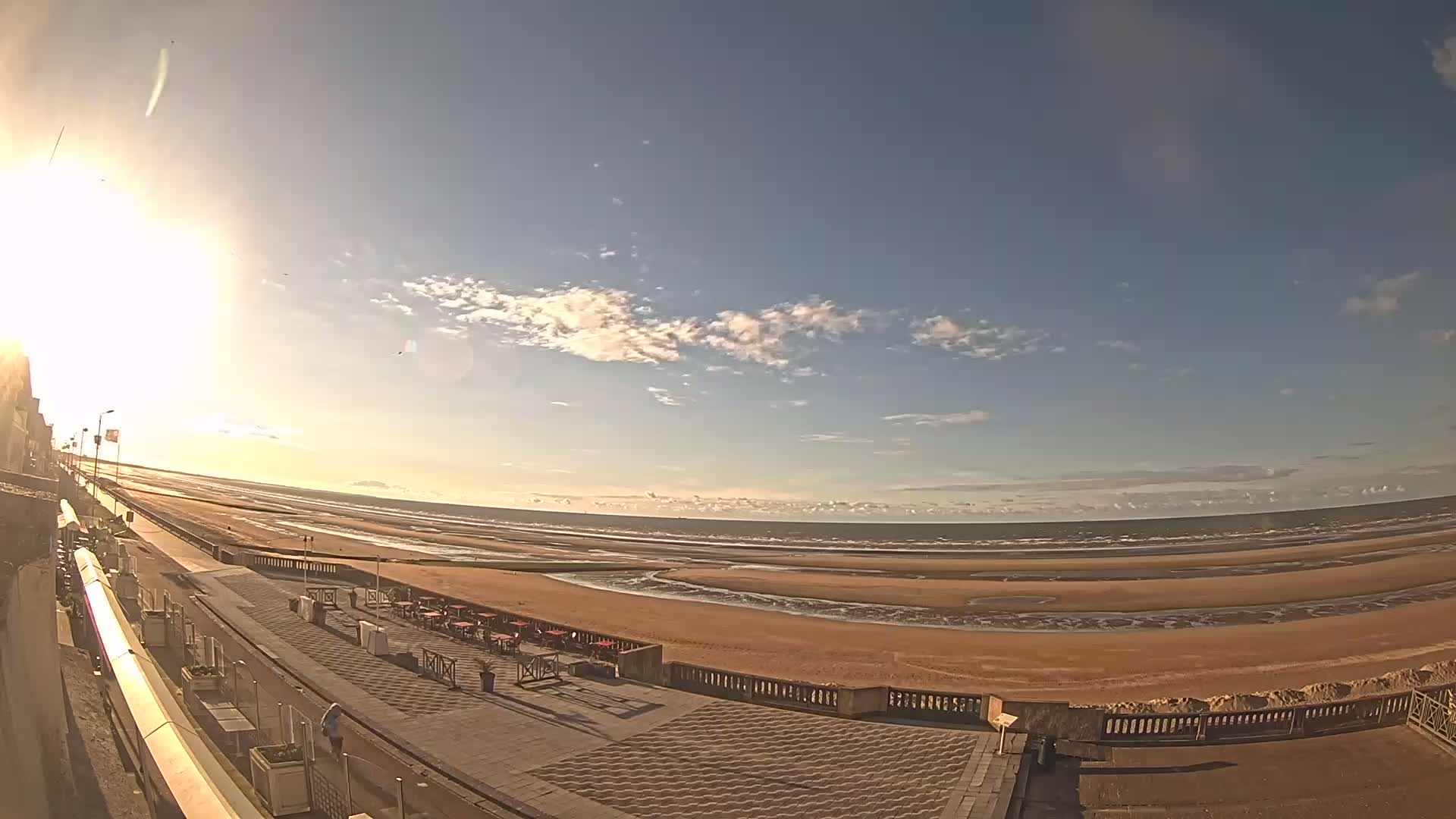 A wide, sunny view of a sandy beach and ocean, with a paved walkway and outdoor seating area in the foreground under a mostly clear sky.