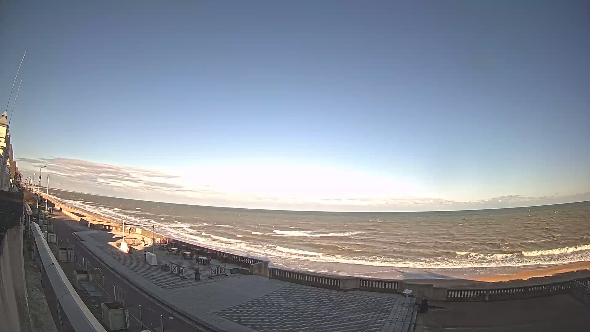 A mostly clear, sunny day shows a wide, wavy ocean meeting a paved beachfront promenade next to a building.