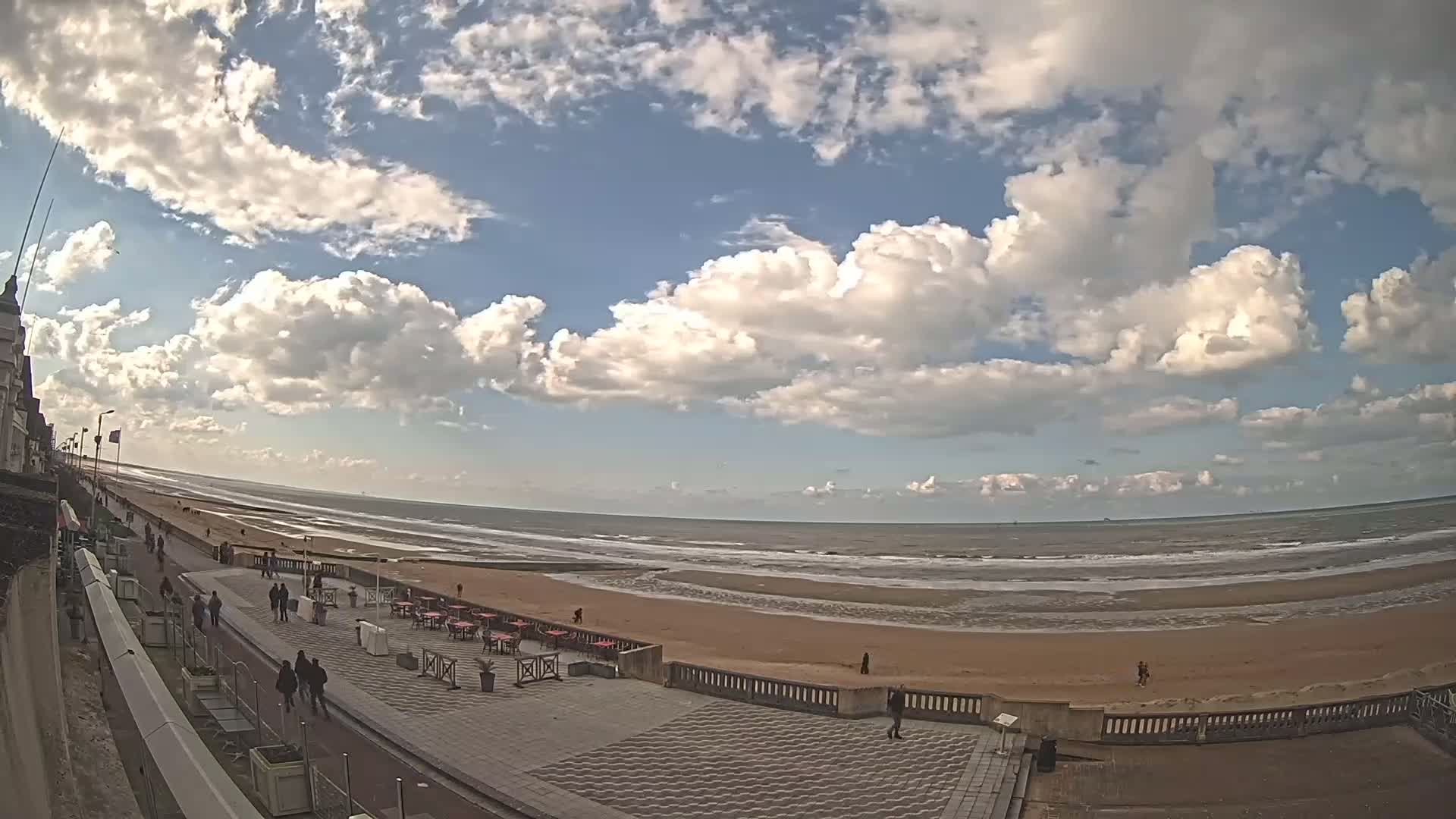 A mostly sunny day with puffy clouds overhead shows a wide sandy beach, ocean waves, and a paved walkway lined with people and outdoor seating.