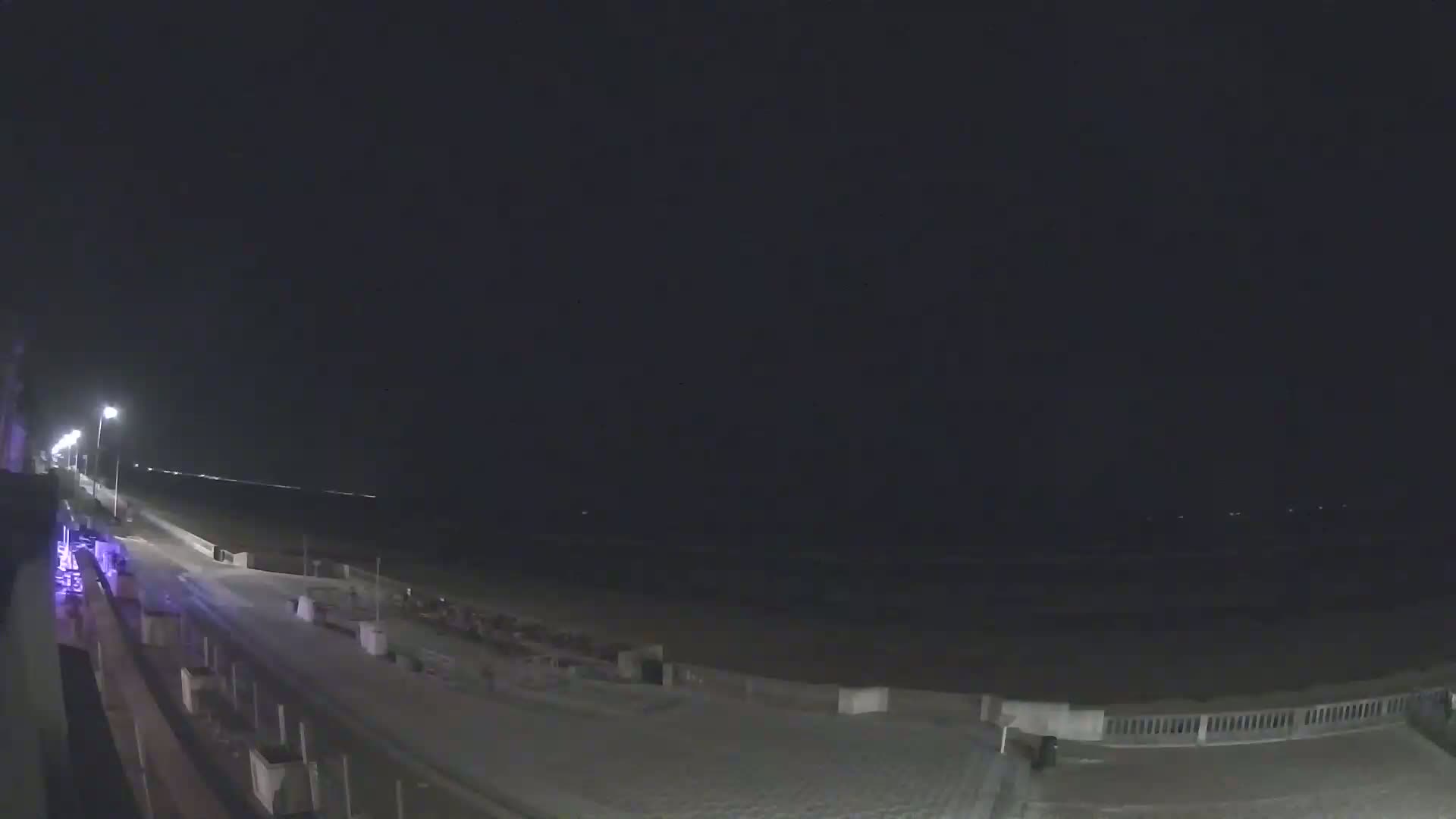 A nighttime view of a mostly empty beach and boardwalk, lit by streetlights, under a dark sky.
