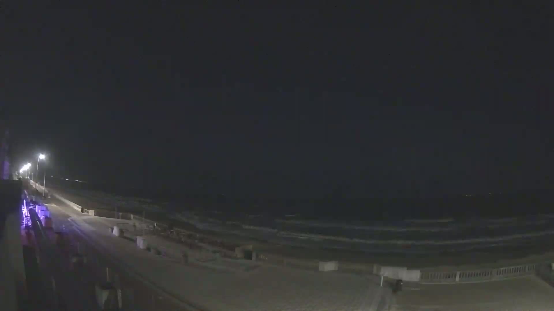 A nighttime view of a beach with gently rolling waves under a dark sky, illuminated by streetlights along a paved walkway.