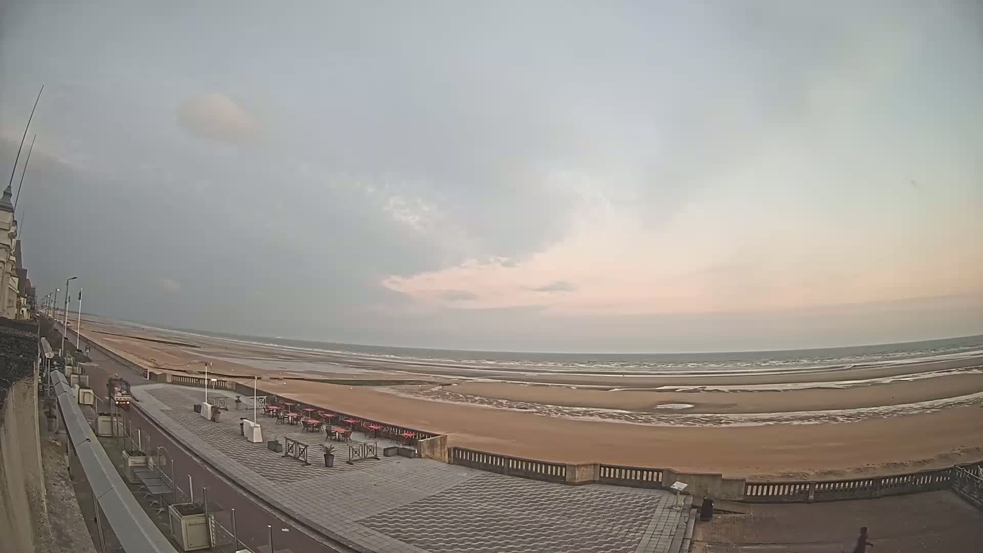 A wide shot of an overcast beach scene shows a paved promenade with outdoor seating next to a wide sandy beach and ocean under a pale, cloudy sky.