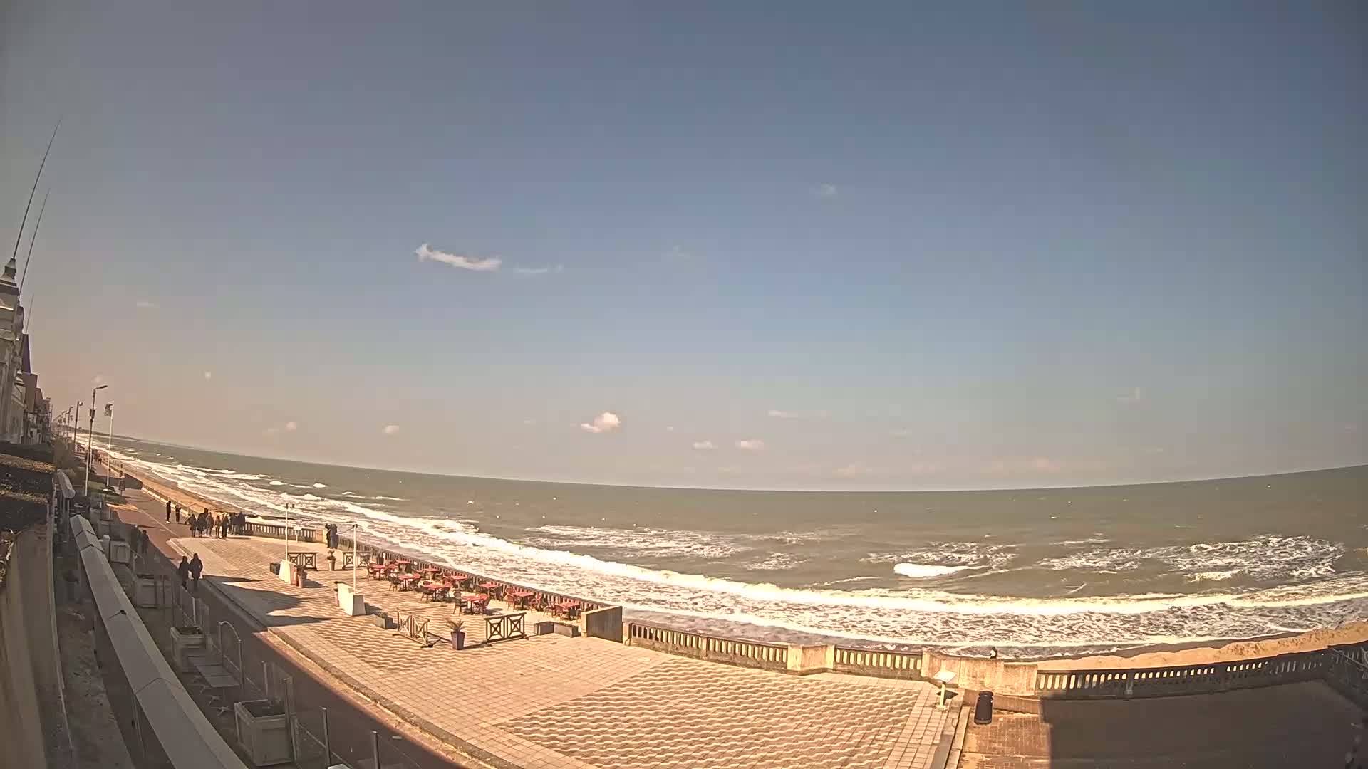 A paved beachfront promenade with outdoor seating overlooks a wavy ocean under a mostly sunny sky.