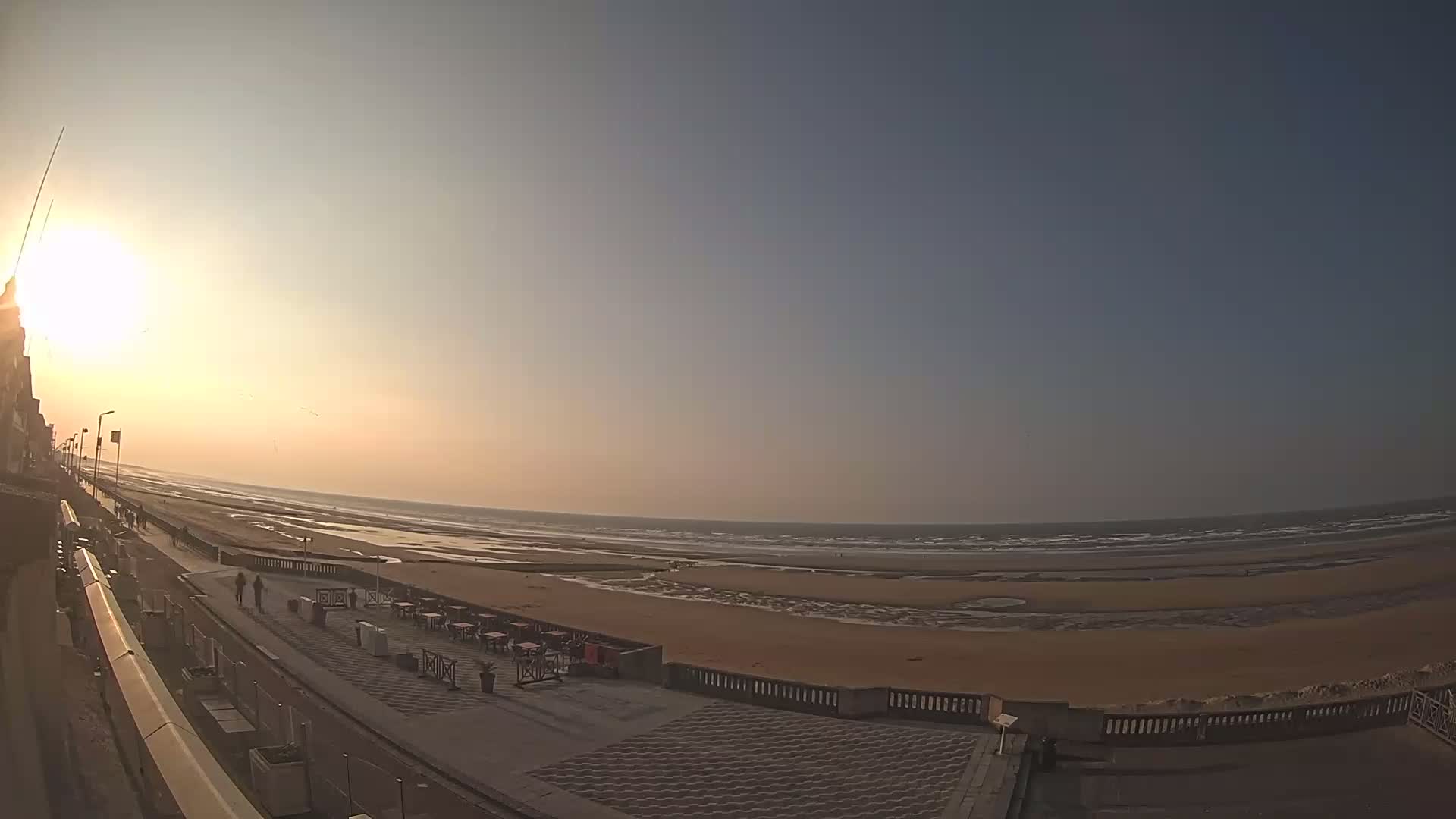 A seaside promenade with a few people walking on it overlooks a wide sandy beach and calm ocean under a bright, sunny sky.