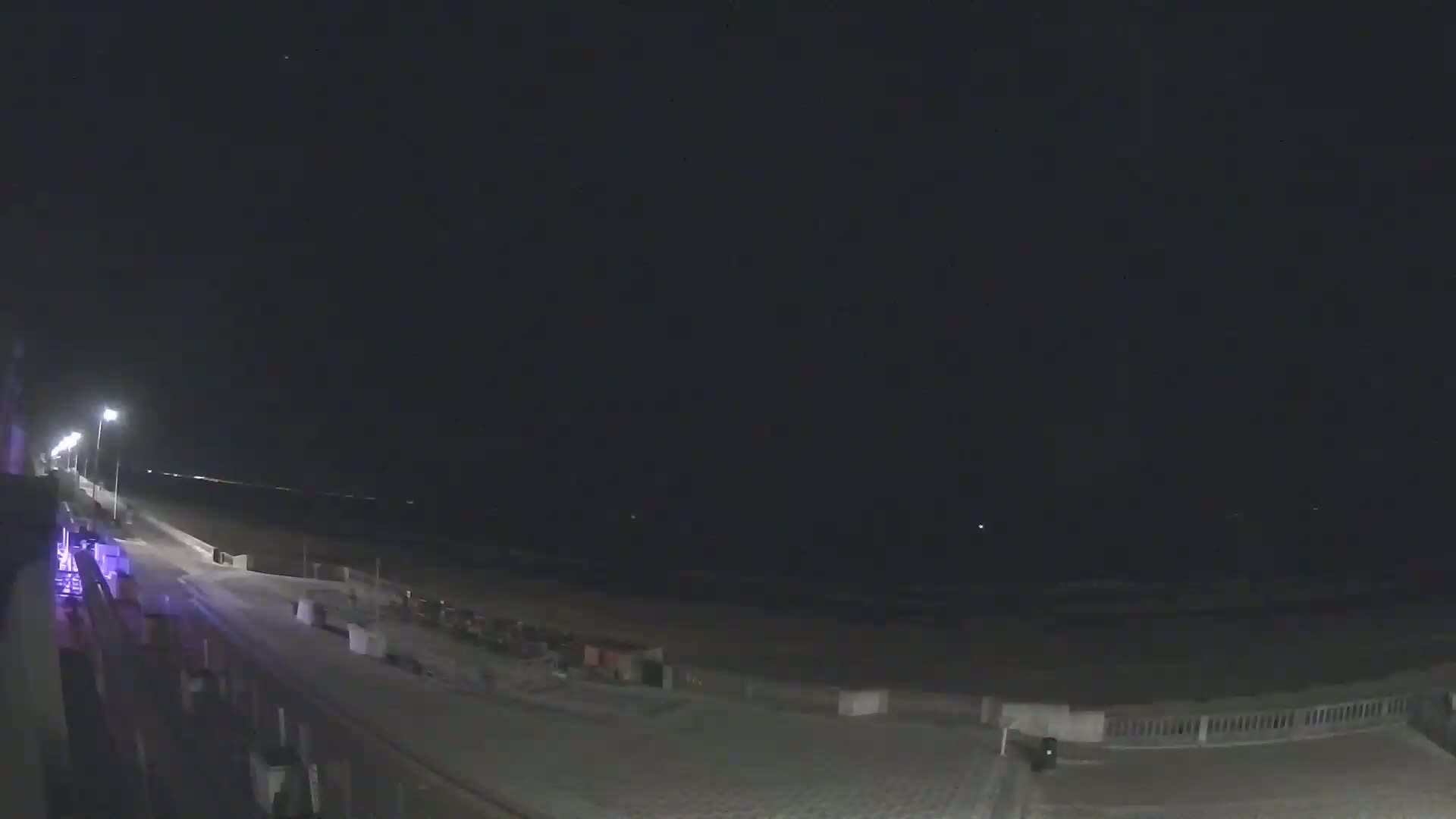 A nighttime view of a beach with a boardwalk and a few lights, under a clear, dark sky.