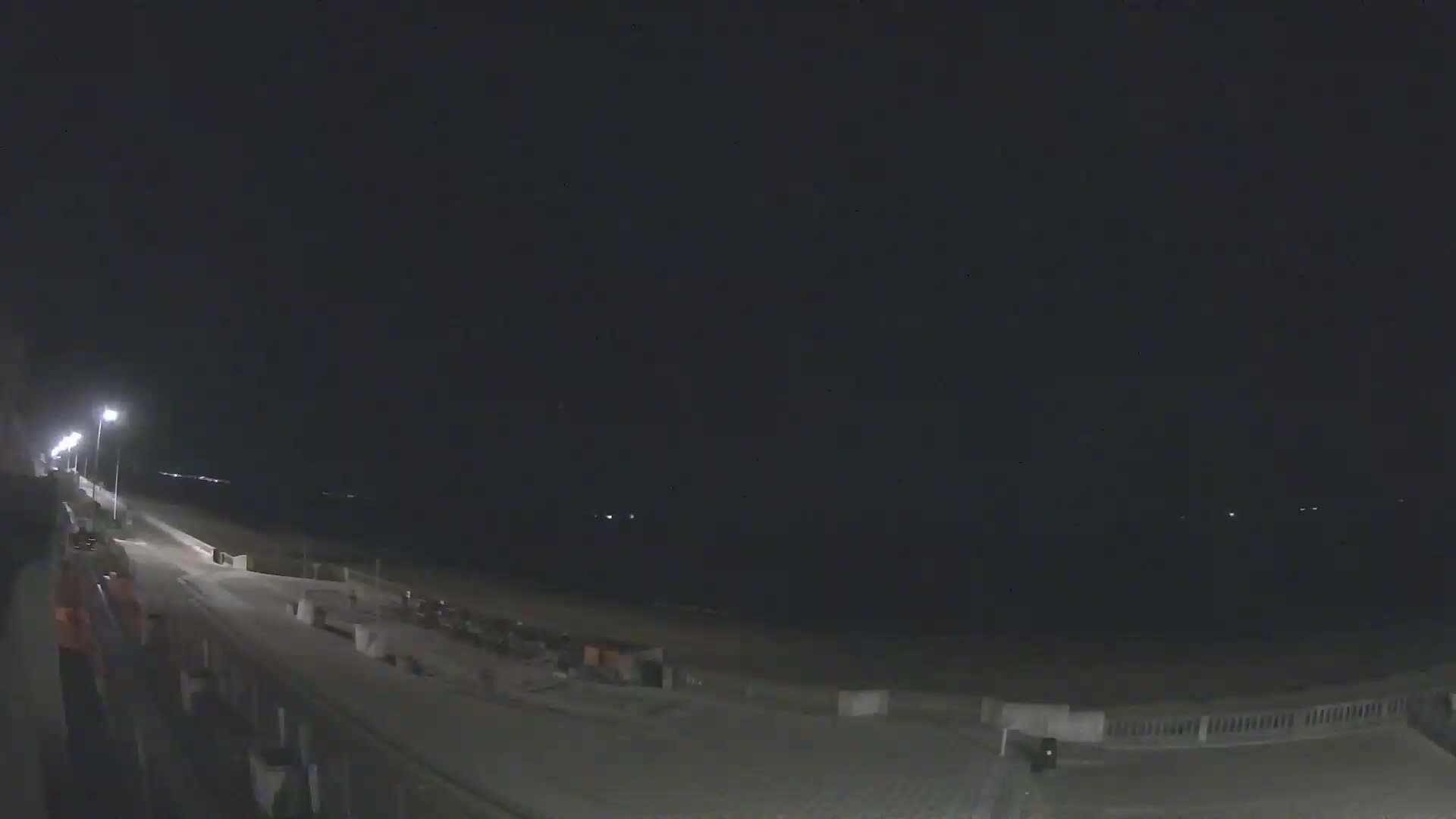 A nighttime view of a mostly empty boardwalk and beach, illuminated by streetlights under a dark sky.