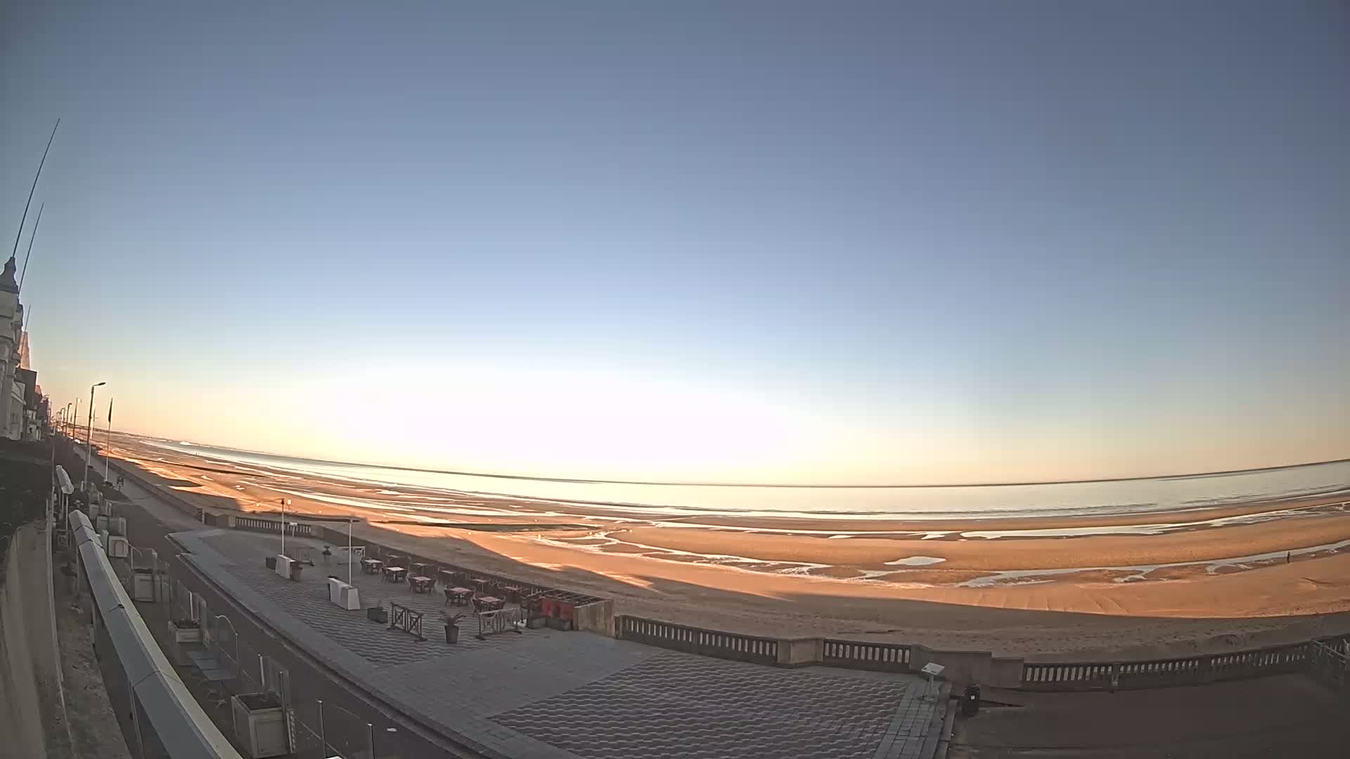 A wide, sandy beach stretches to a calm sea under a clear, bright sky at sunrise, with a paved promenade and outdoor seating area in the foreground.